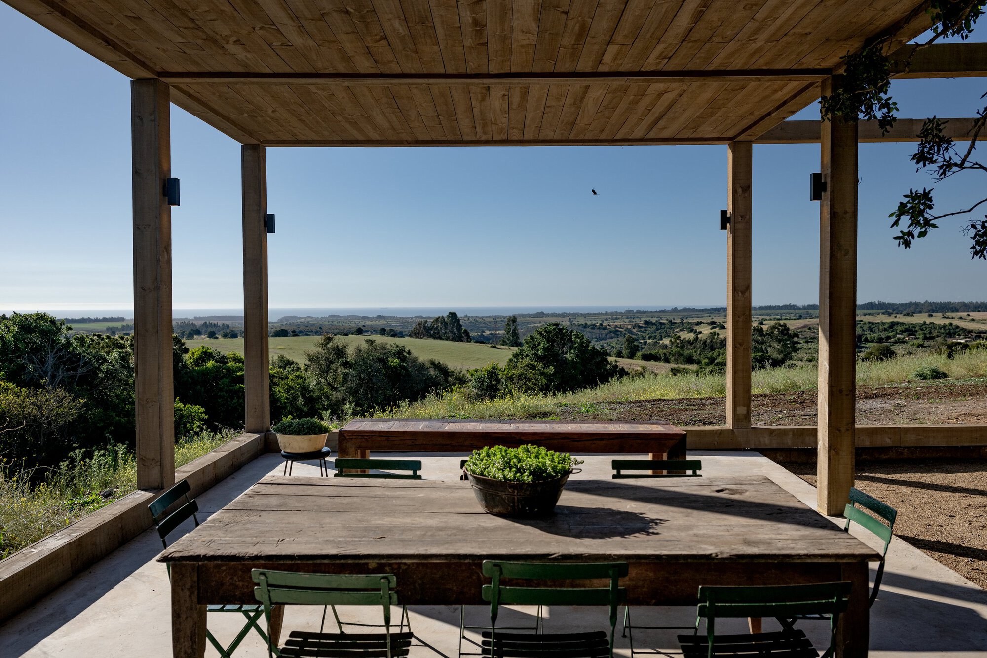 Covered terrace with timber posts and plank ceiling, a rustic dining table and green chairs facing rolling hills and distant ocean