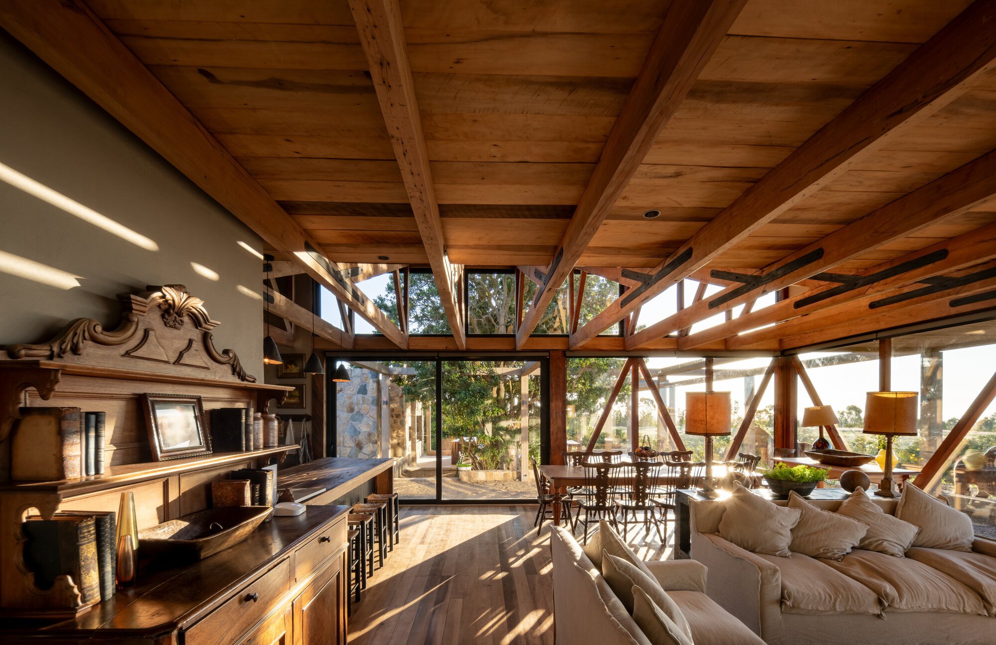 Living room with exposed timber beams and diagonal bracing, an antique carved sideboard and sofa, lit by low raking sunlight through glazing