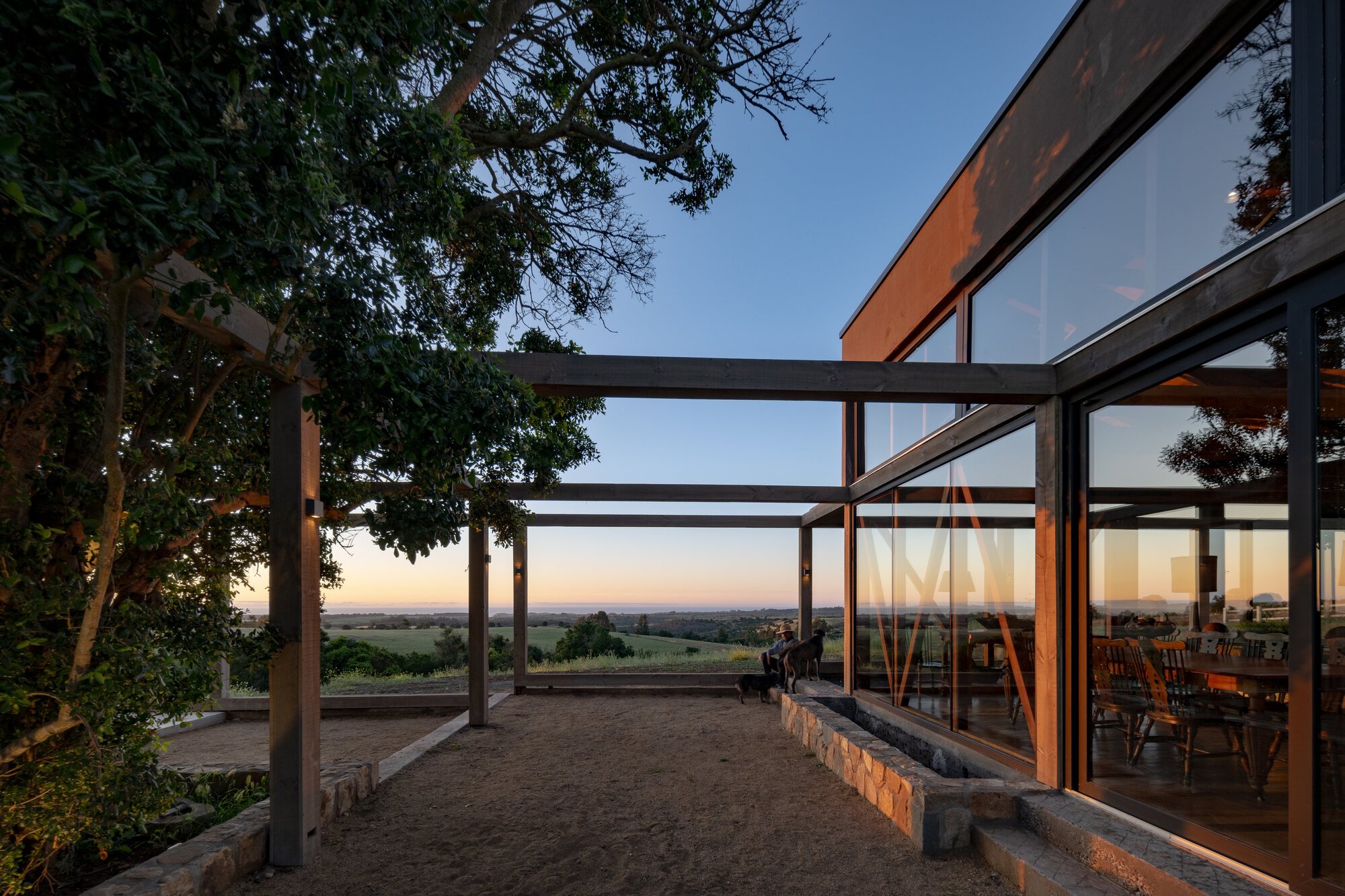 Gravel courtyard at dusk beside a glazed dining room, with a timber pergola, stone planter edge and an oak tree against an orange horizon