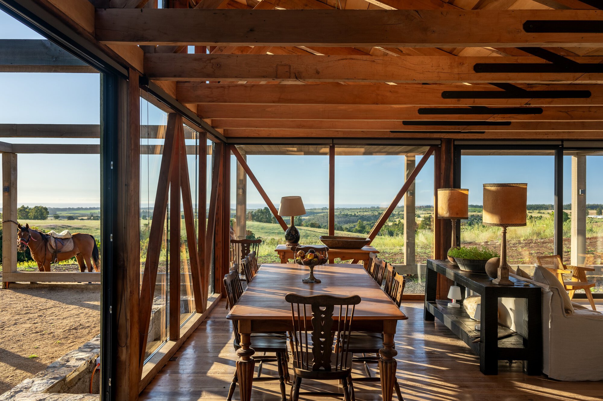 Dining table beneath exposed timber beams and diagonal bracing, with a saddled horse visible through glass and rolling fields beyond