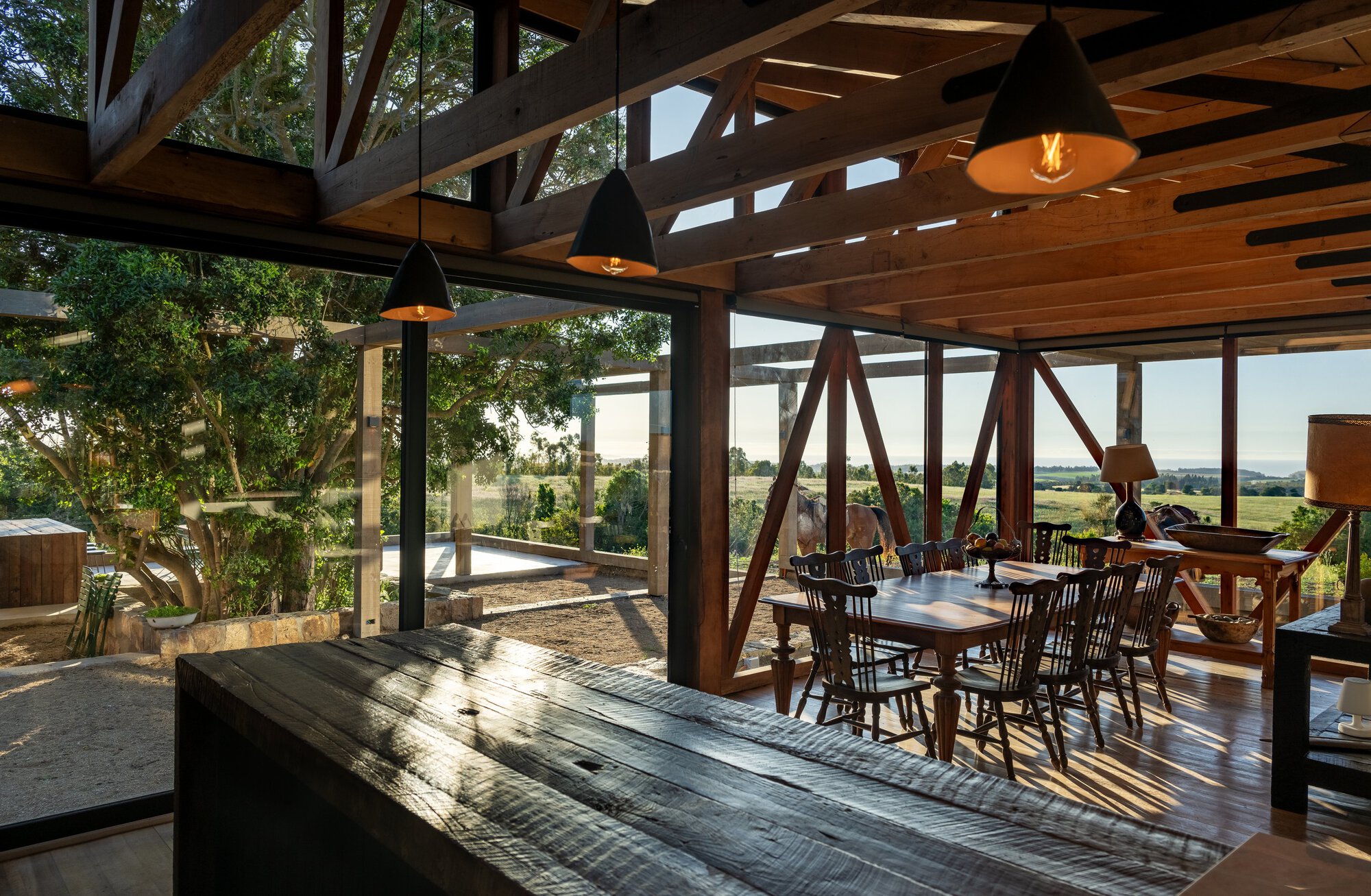 Dining area seen past a reclaimed wood counter, with black pendant lamps, braced timber frame and late sun streaking across the floor