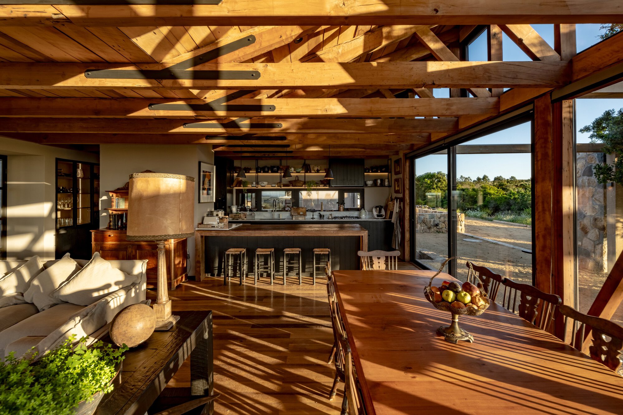 Open living space with sofa, kitchen island and bar stools under sunlit timber rafters, a fruit bowl on the dining table