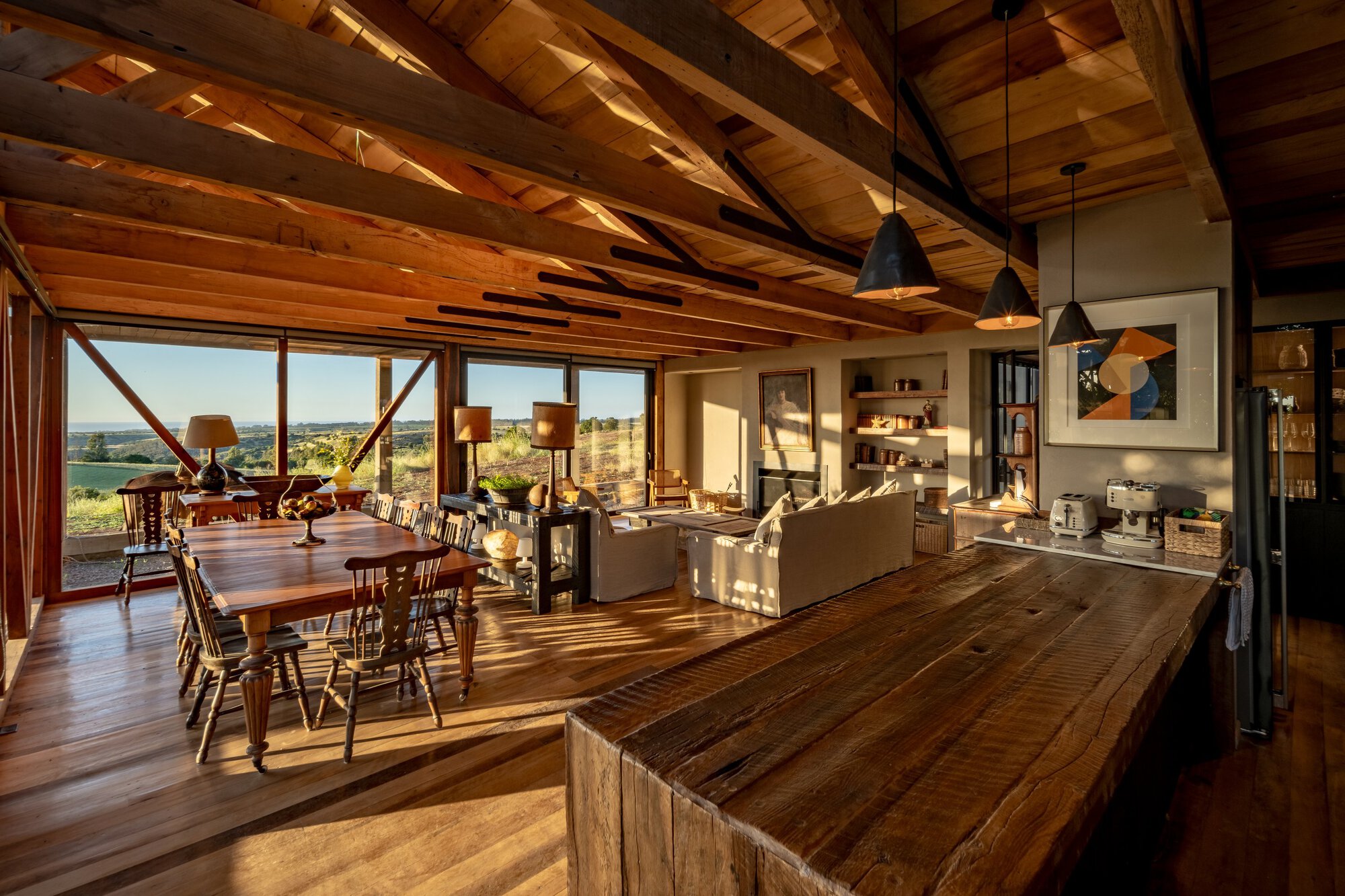 Open living and dining room with exposed timber trusses and a reclaimed wood island, lit by low golden sunset light