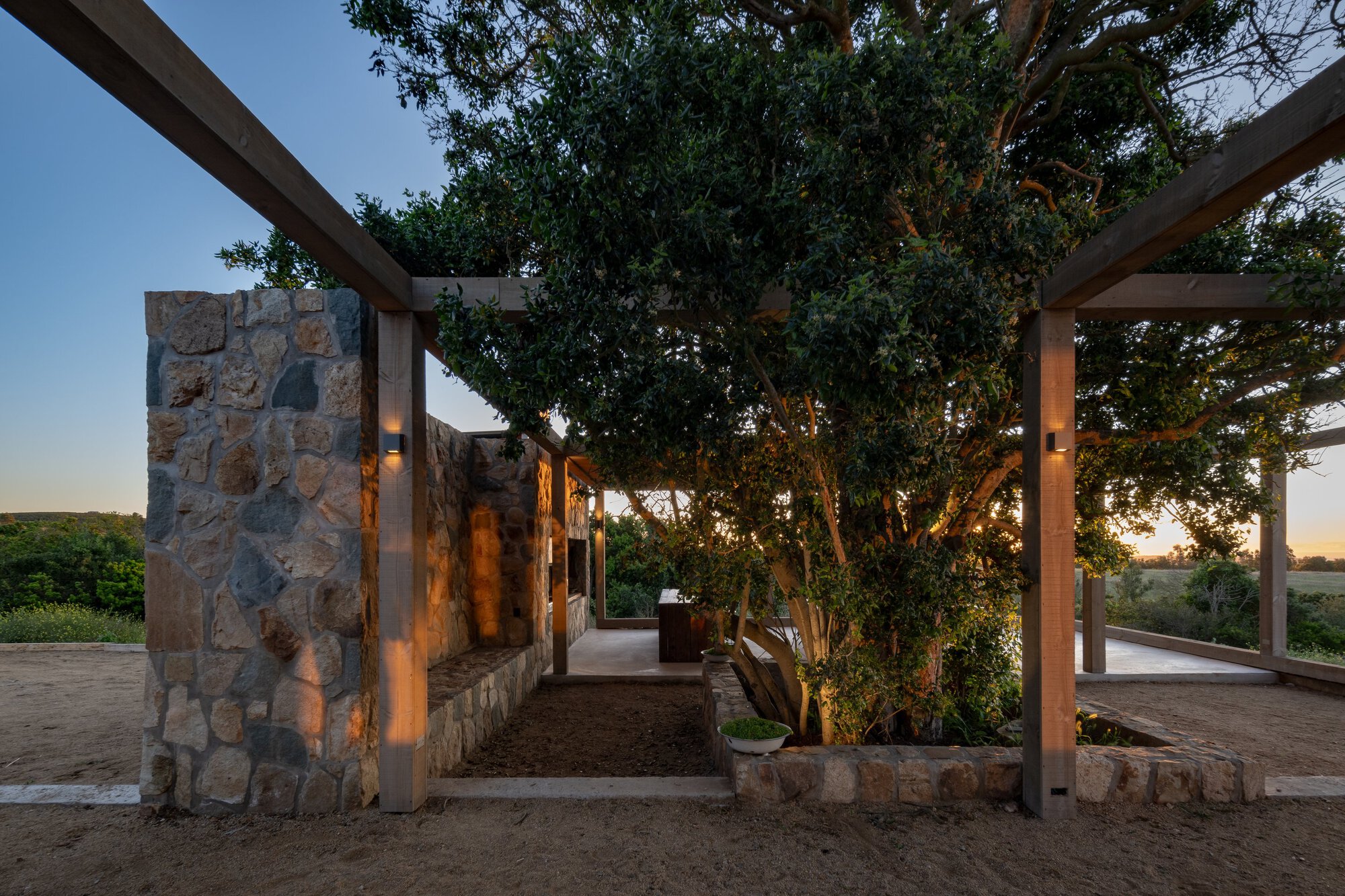 Stone wall and timber posts with wall lights framing a mature tree in a gravel courtyard at twilight