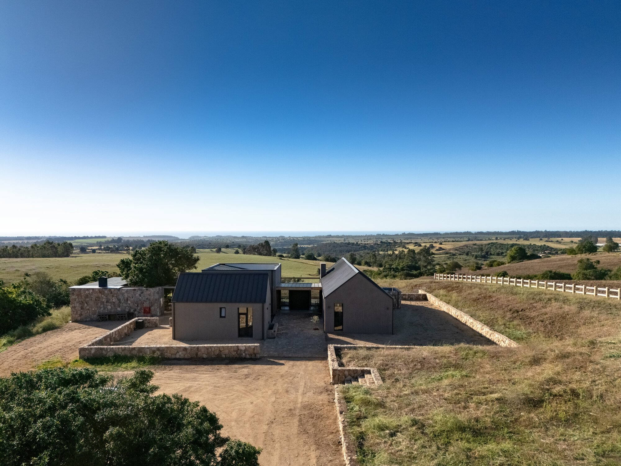 Drone view of the gabled dark-roofed house with stone retaining walls on a grassy hillside under a clear blue sky