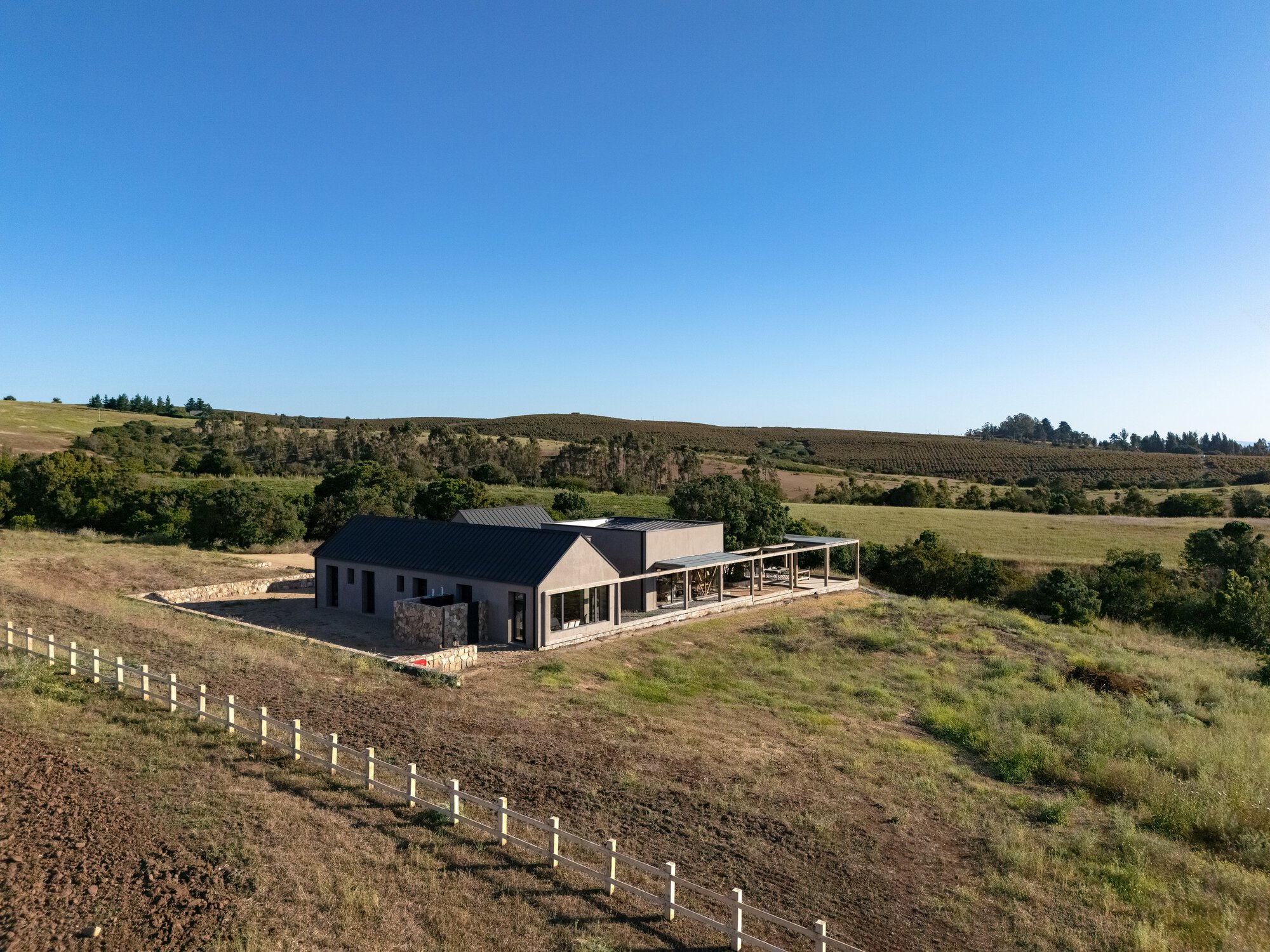 Aerial view of the low gabled house and covered timber pergola beside a white fence in rolling farmland