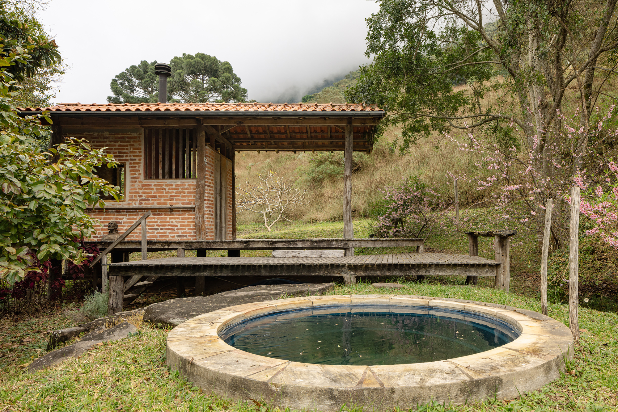 Round stone-rimmed pool in front of a brick cabin with raised timber deck, under low cloud and blossoming trees