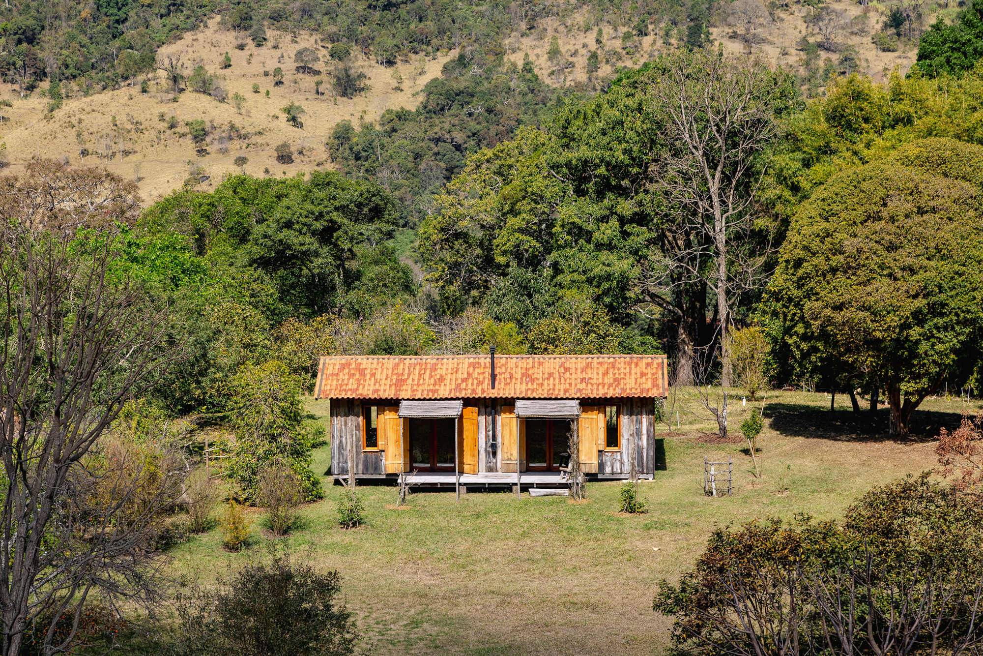 Weathered timber cabin with tiled roof and yellow shutters on a grass clearing, backed by green trees and dry hillside