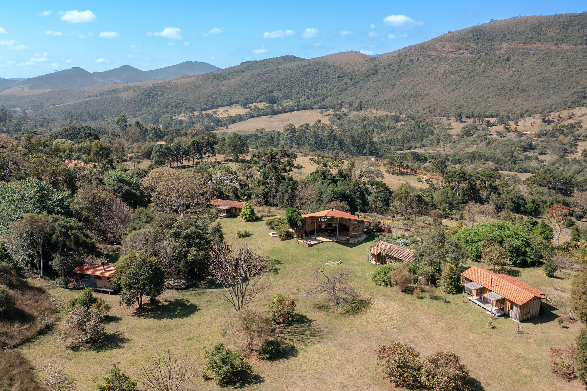 Aerial view of several small tile-roofed buildings scattered across a grassy clearing, with forested hills beyond under a blue sky