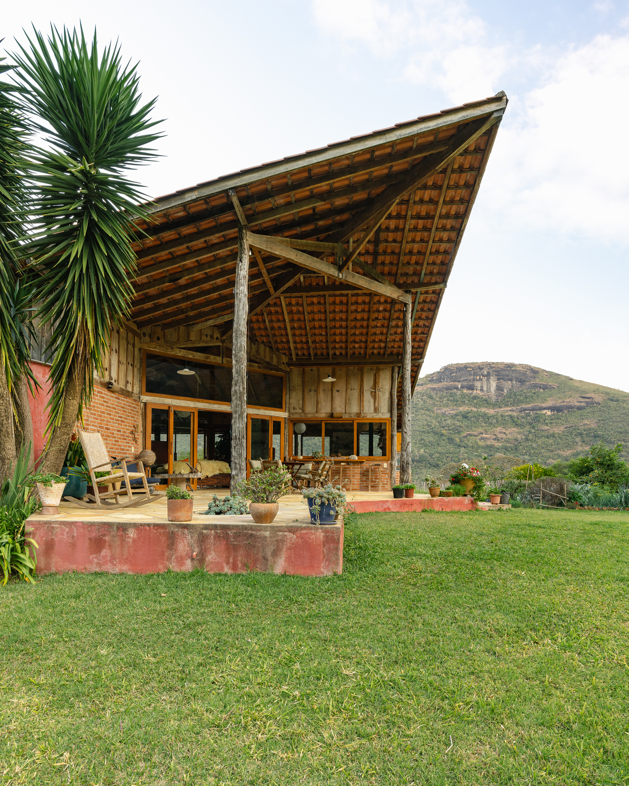 Steeply pitched clay-tile roof on timber posts over a brick and glass house, with a yucca and green lawn in front