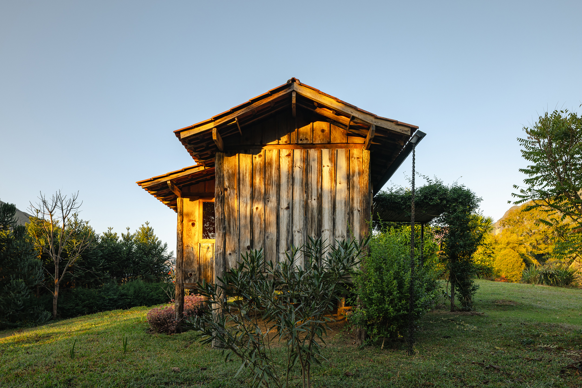 Gabled end of a vertical board cabin with layered eaves, framed by shrubs and a hanging chain at dusk