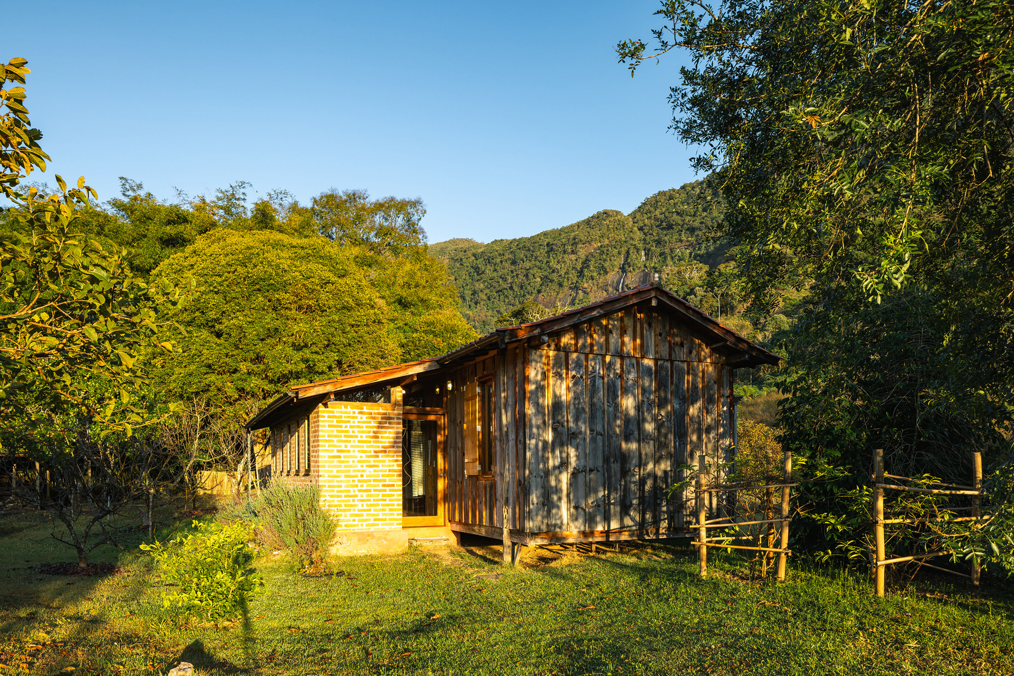 Brick entry volume joined to a board-and-batten cabin, with a rustic wooden fence and dappled shadows from nearby trees