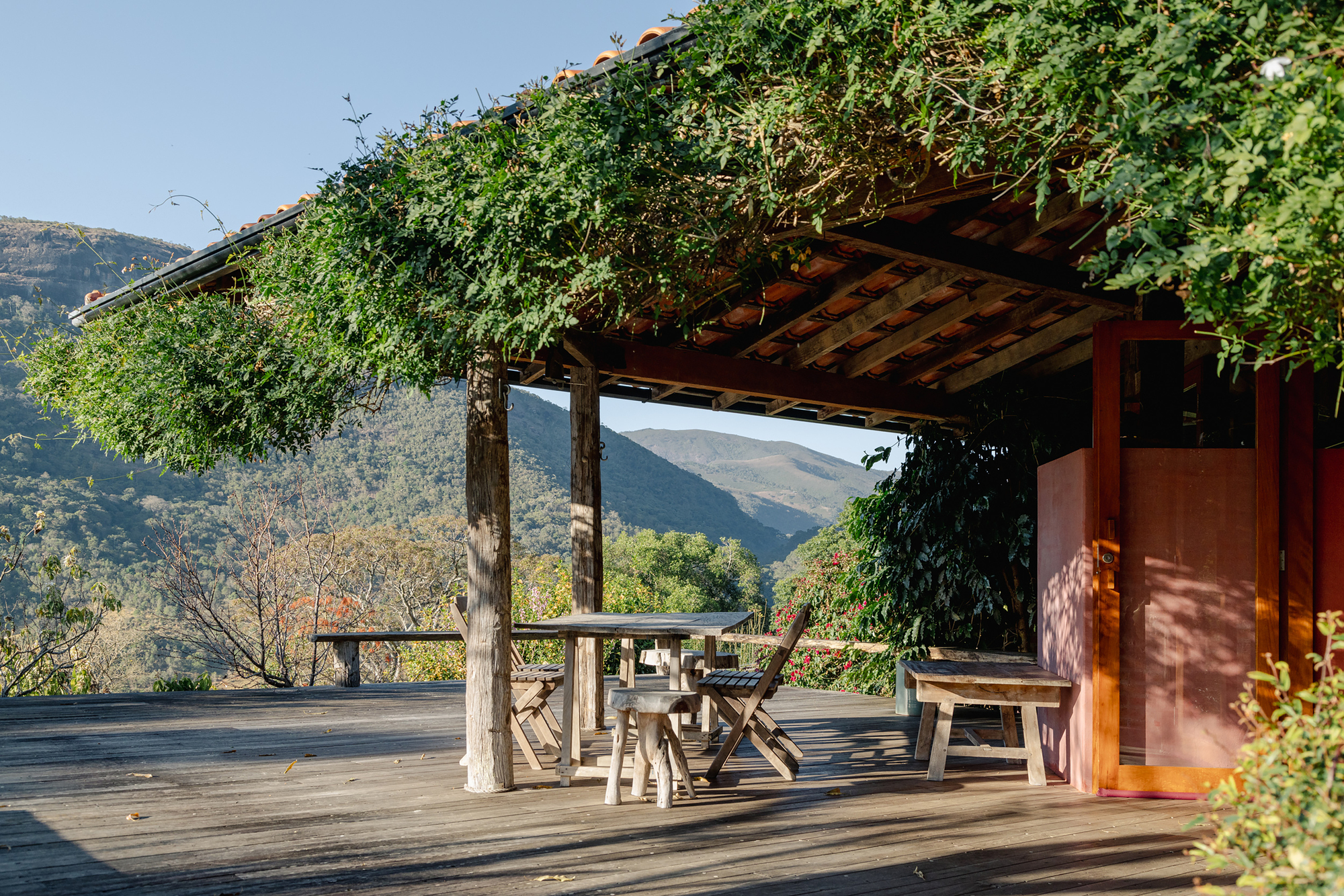 Covered timber deck with rustic table and folding chairs under a vine-draped roof, mountains behind in clear daylight
