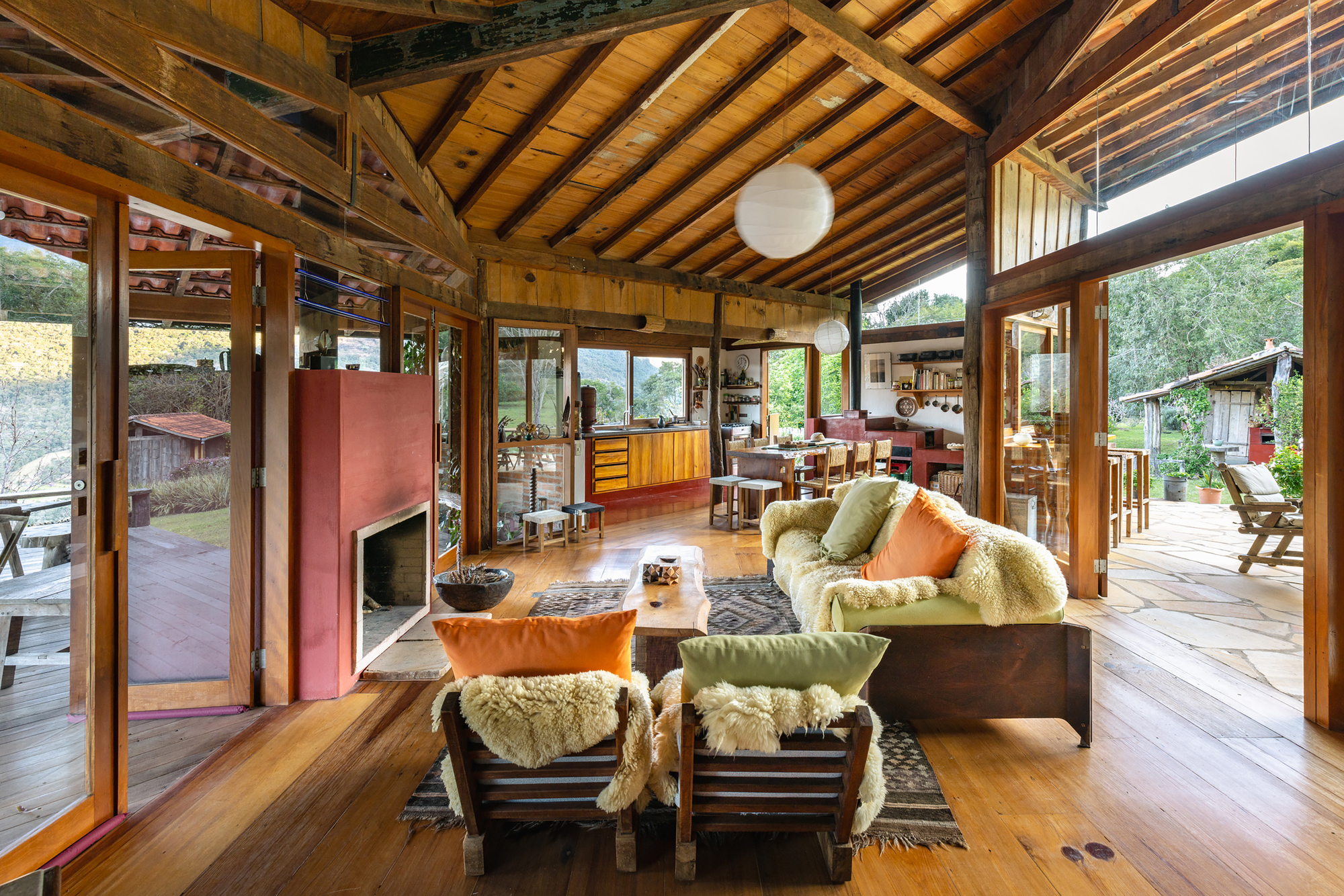 Open living room with red fireplace volume, sheepskin-covered sofas and timber ceiling, glazed doors opening to a stone terrace