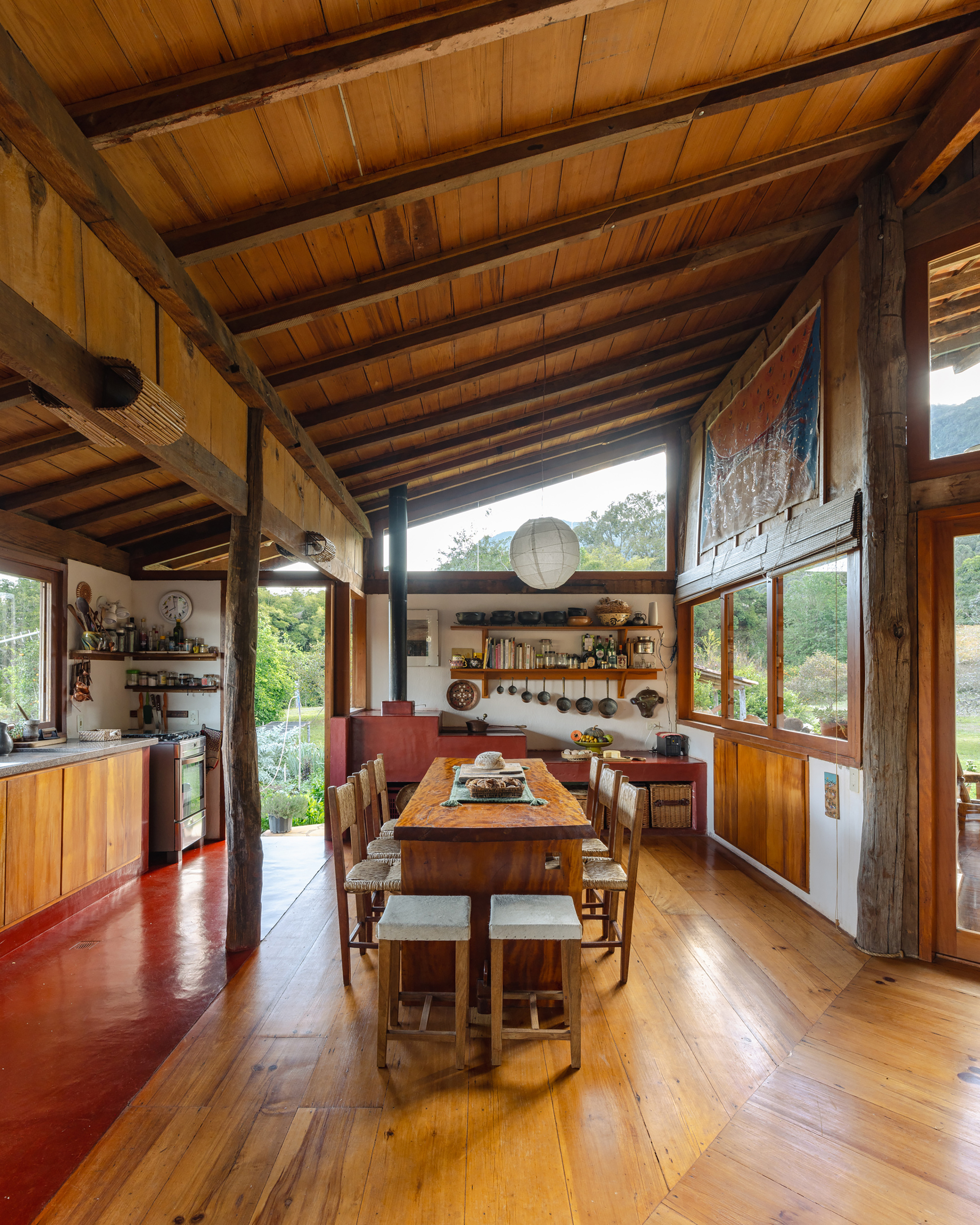 Kitchen and long wooden dining table beneath sloped timber ceiling, with red concrete floor and open shelves of cookware