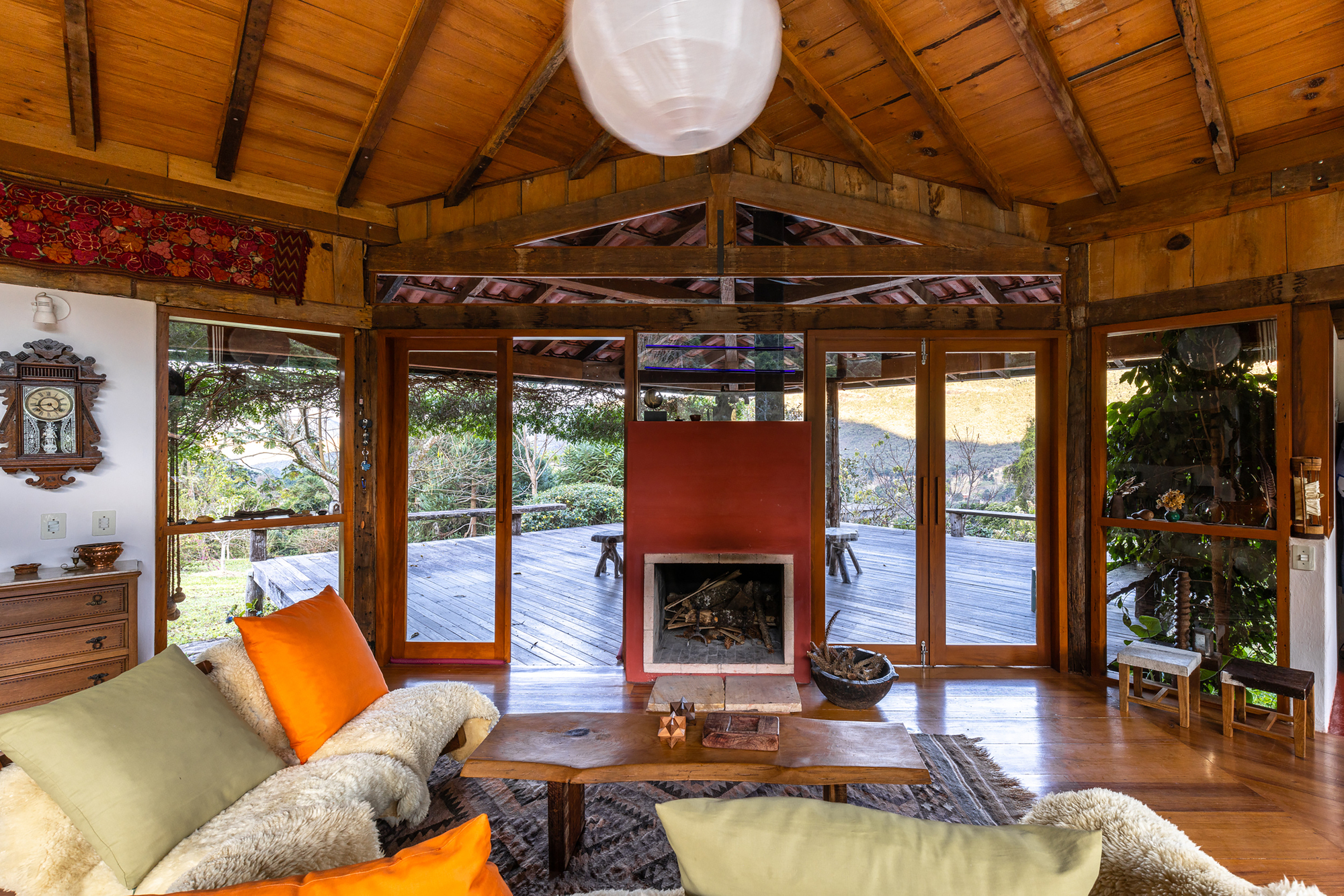 Red fireplace block between glazed doors, framing a wooden deck and hillside beyond, with orange cushions in the foreground