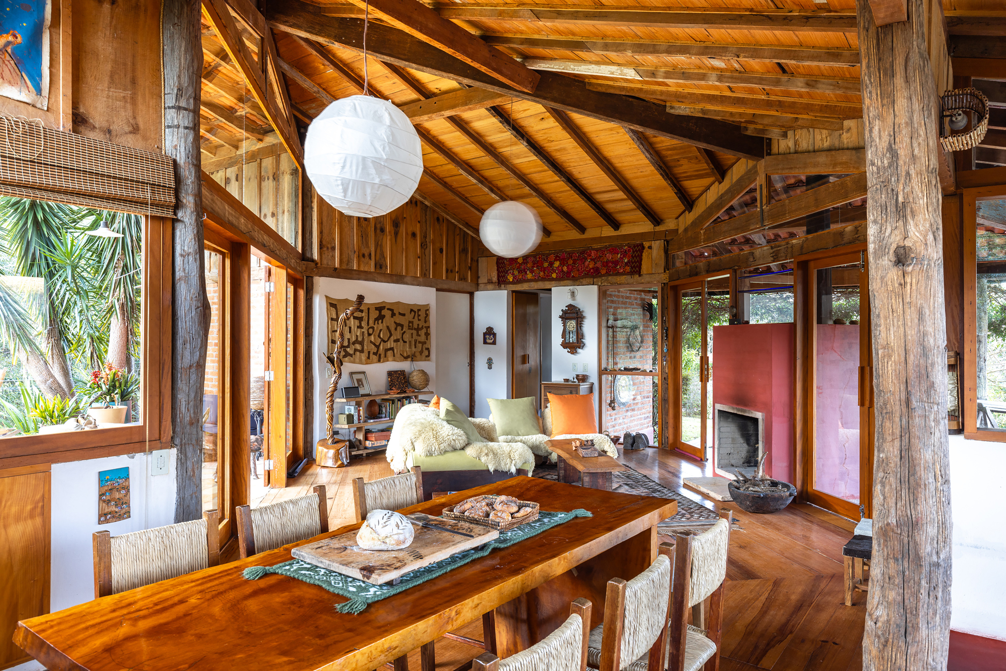 Dining table in foreground with rough timber posts, paper lantern lamps and sloped plank ceiling over the living area