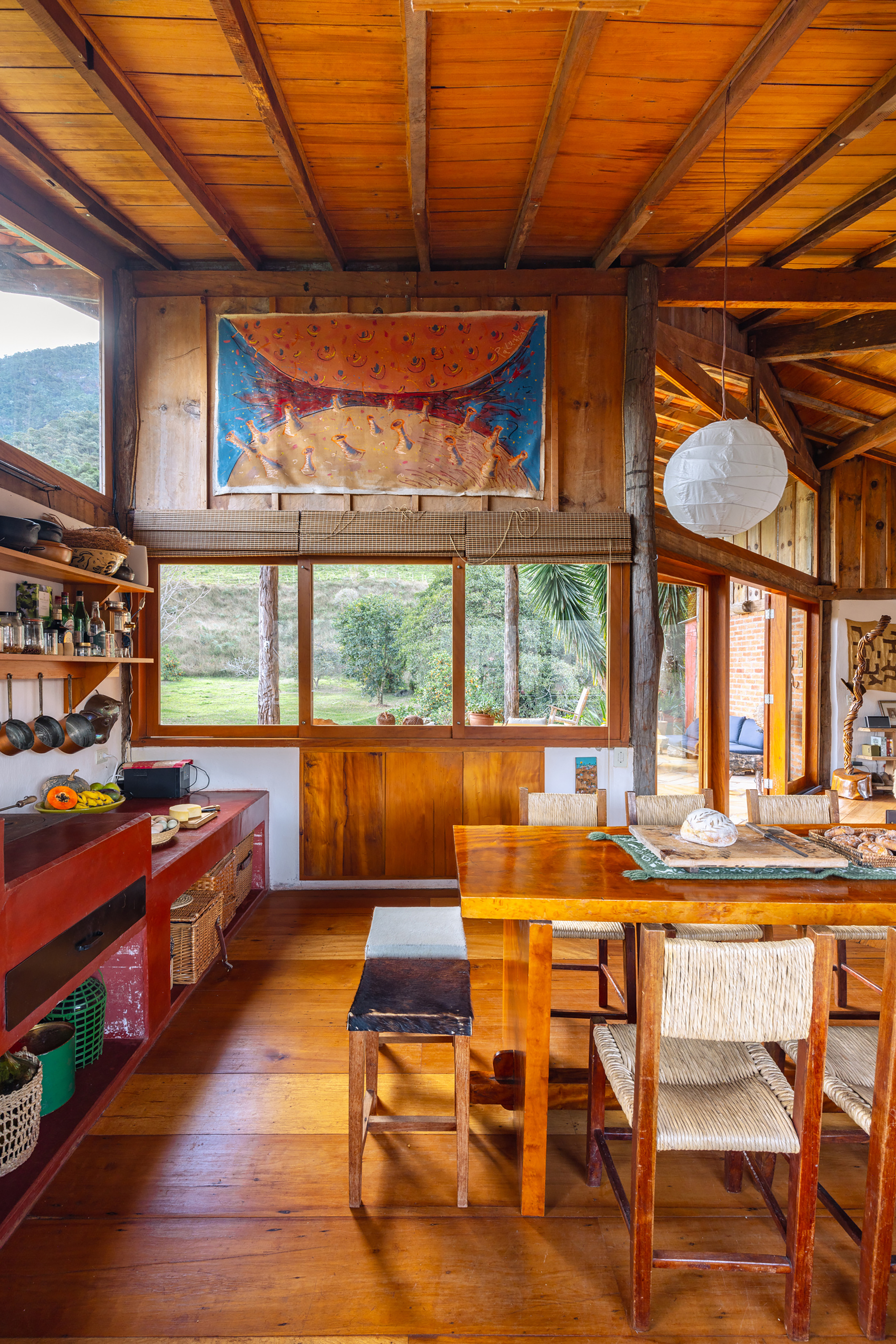 Kitchen corner with red shelving, a large painting above a window and woven chairs at the dining table in daylight