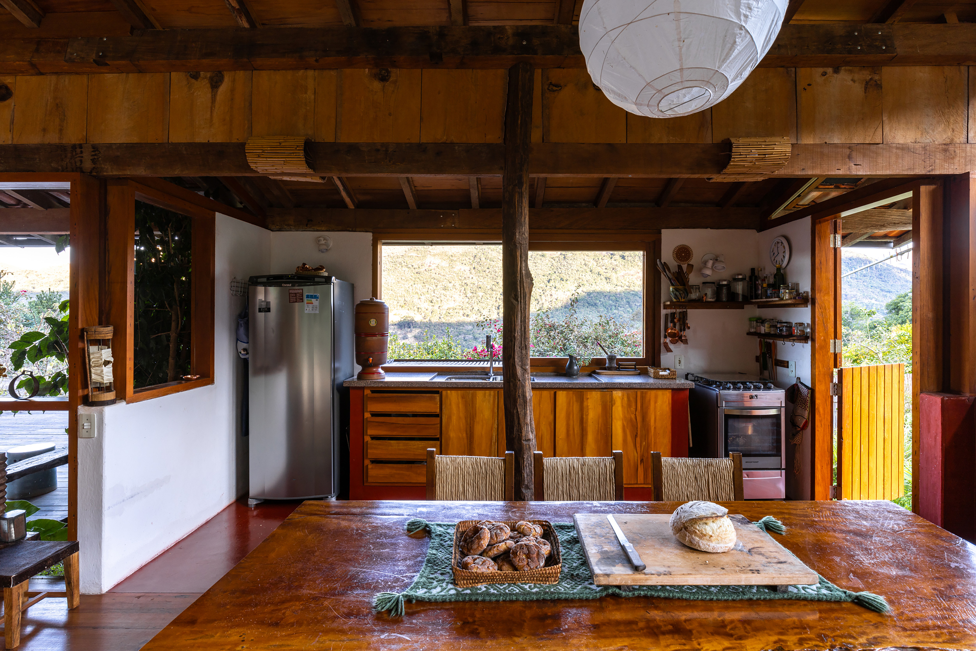 Kitchen and long timber dining table framed by a central post, with window views of green hills in daylight