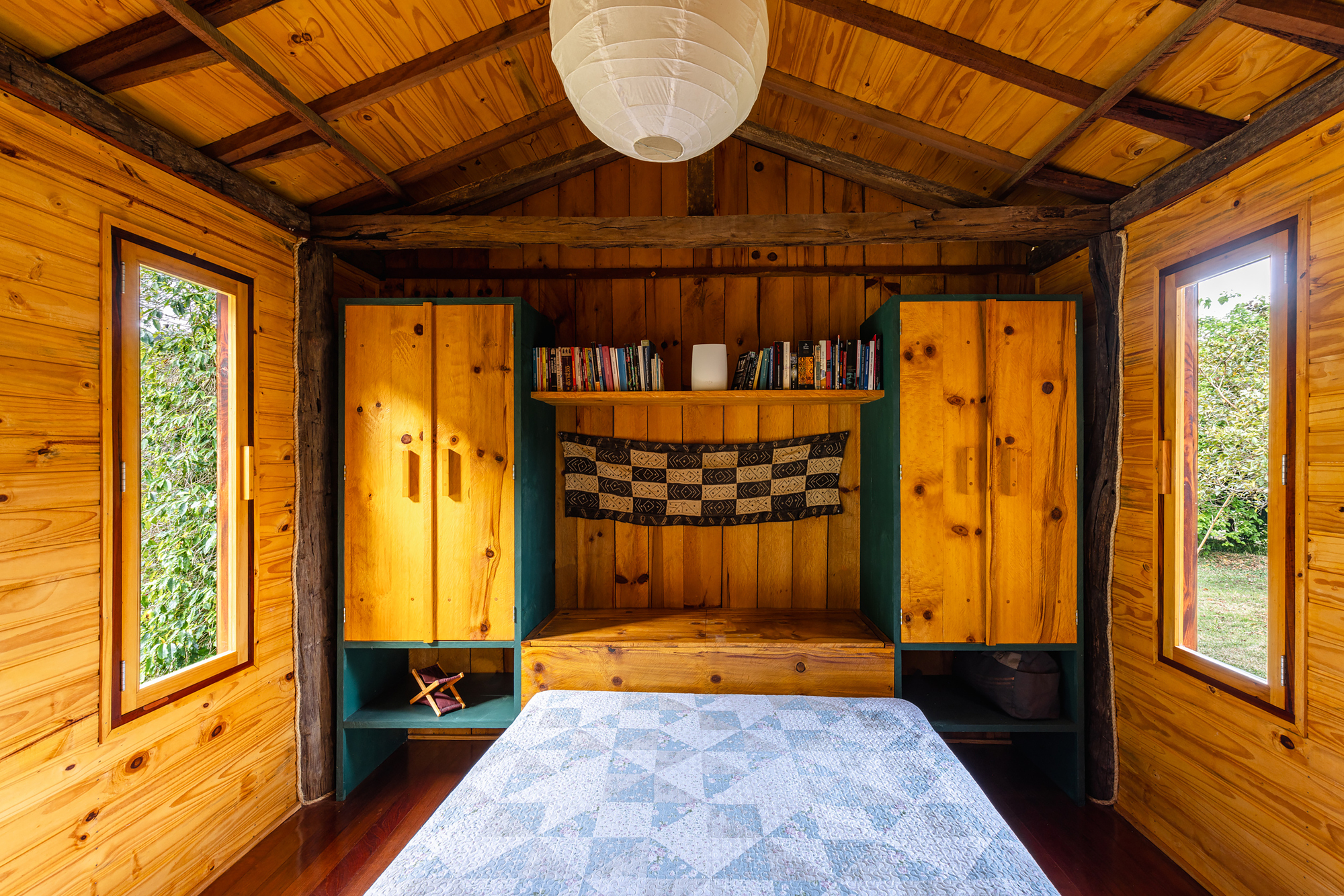 Bedroom with pine-clad walls, built-in teal cabinets and a checkered textile above the bed, with leafy light through narrow windows