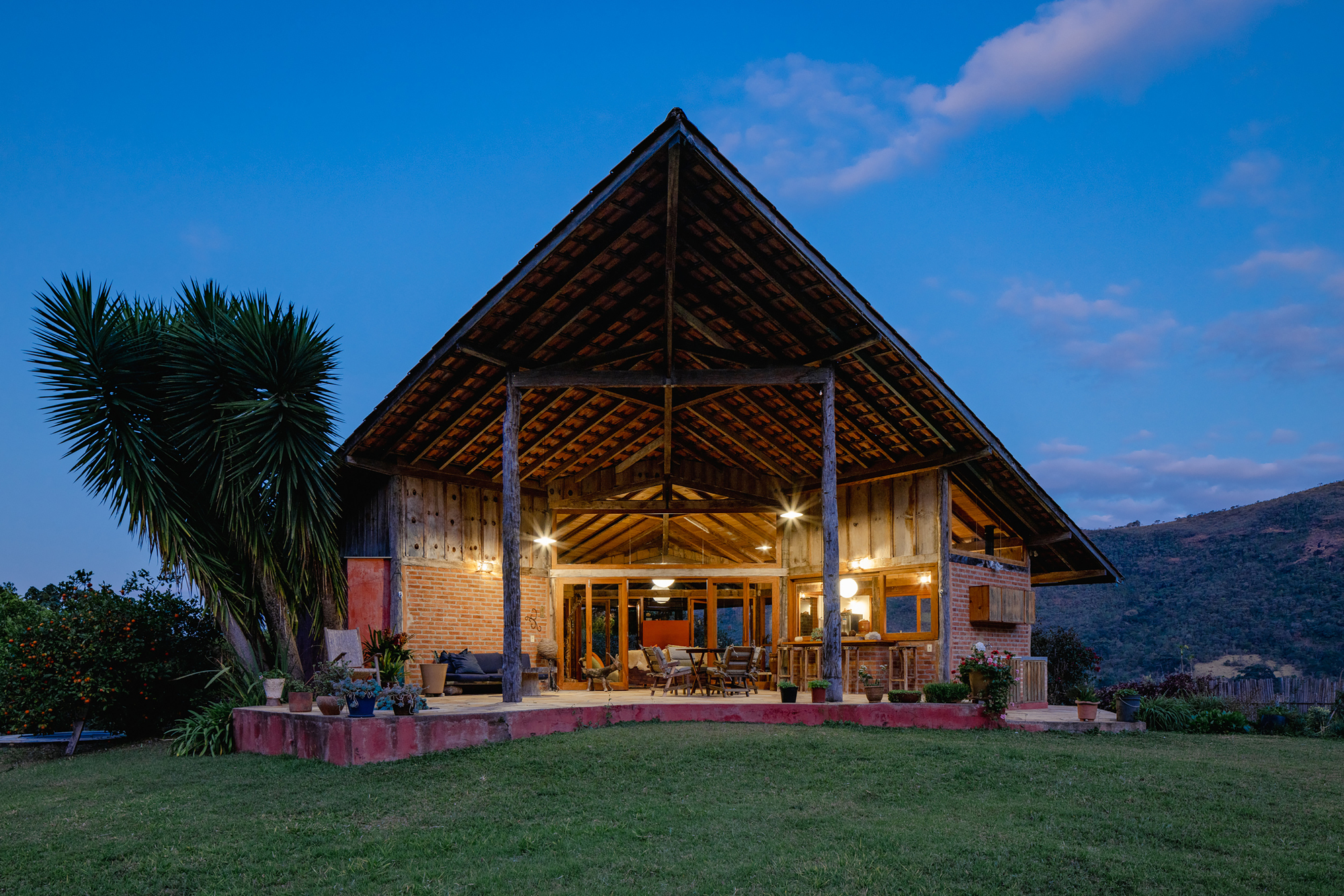 Gabled timber-framed house with open porch and exposed rafters glowing at dusk beside a palm and lawn