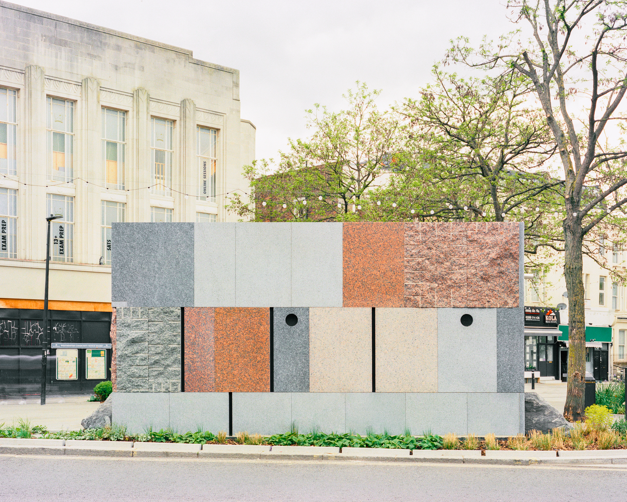 Street-facing wall of the pavilion, built from panels of grey, pink and red granite with two round openings, under an overcast sky