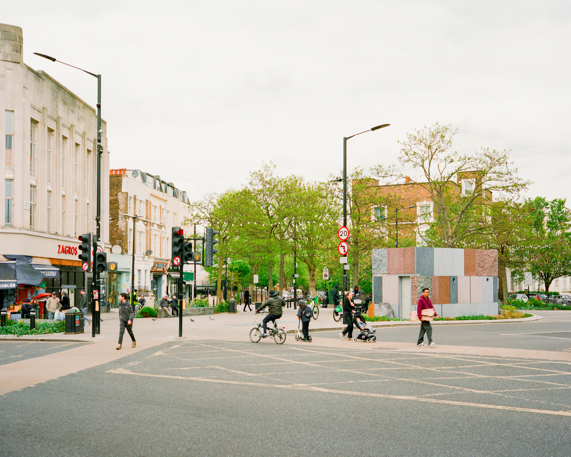 Wide view across a junction with the stone-clad pavilion on a planted island, cyclists and pedestrians crossing in soft grey light