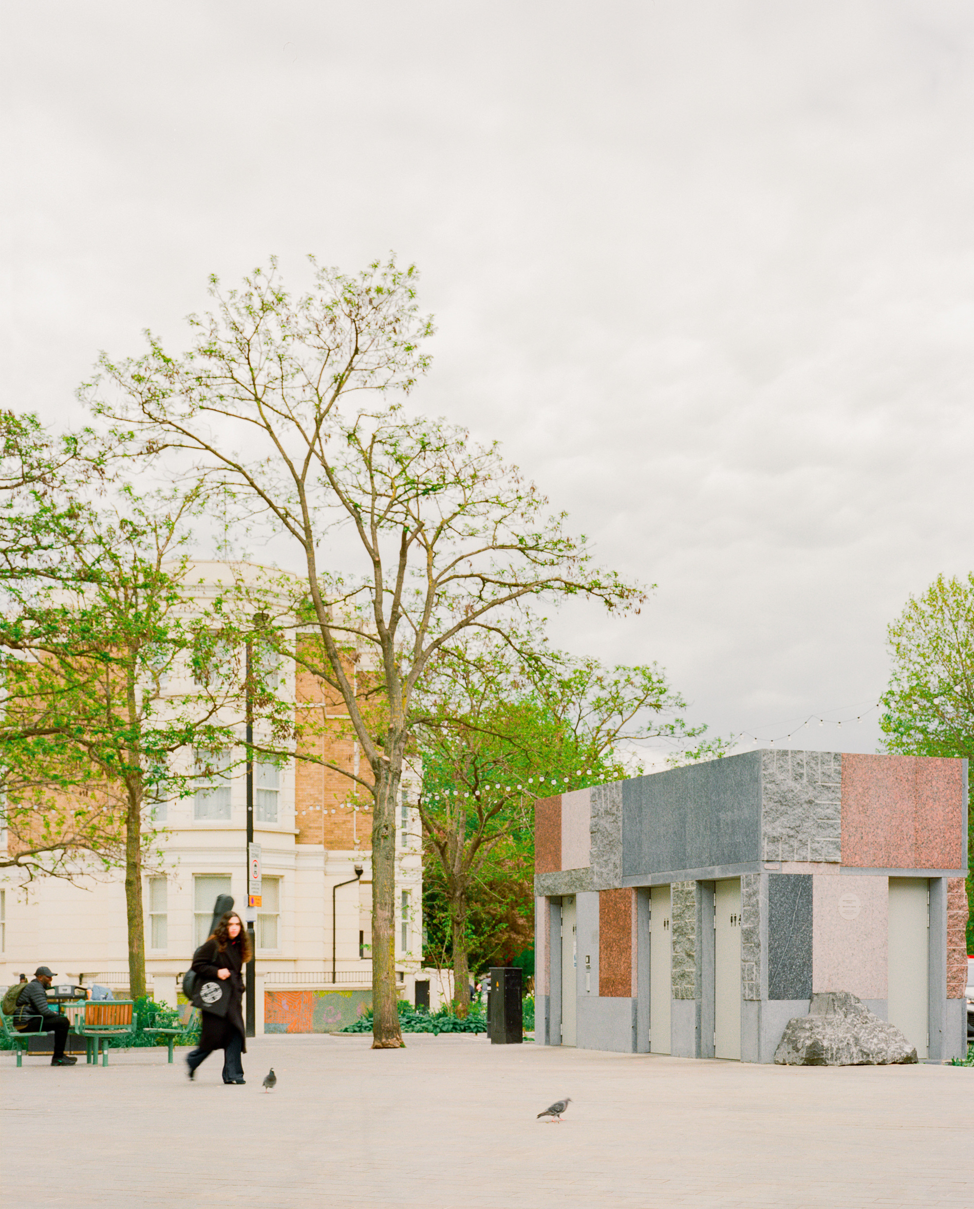Side of the granite pavilion with recessed doors and a boulder at its base, a woman walking past a tall tree