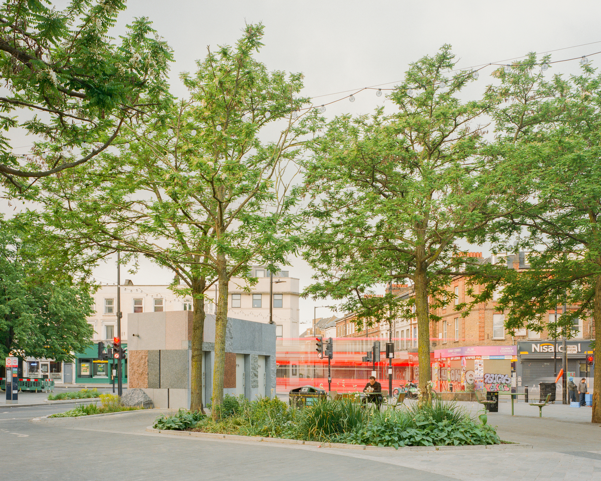 Pavilion partly hidden behind slender trees and a planted bed, a red bus streaking past in the background