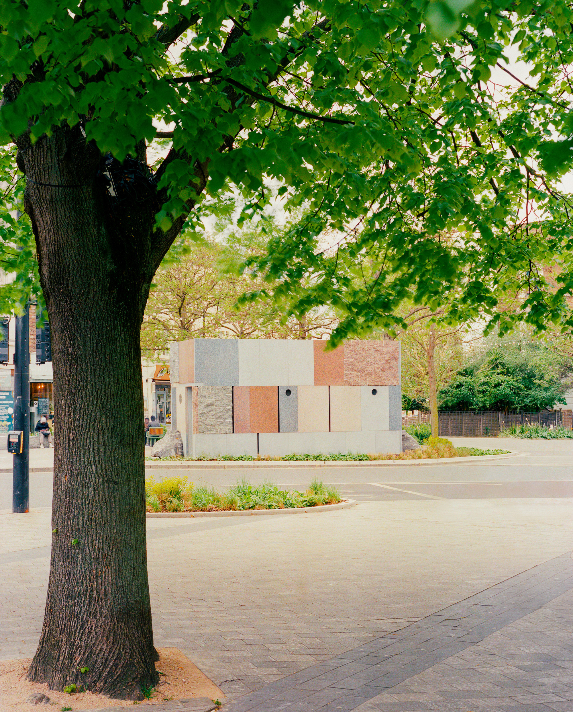 Small pavilion clad in multicoloured stone panels seen across a paved square beneath a leafy tree in spring