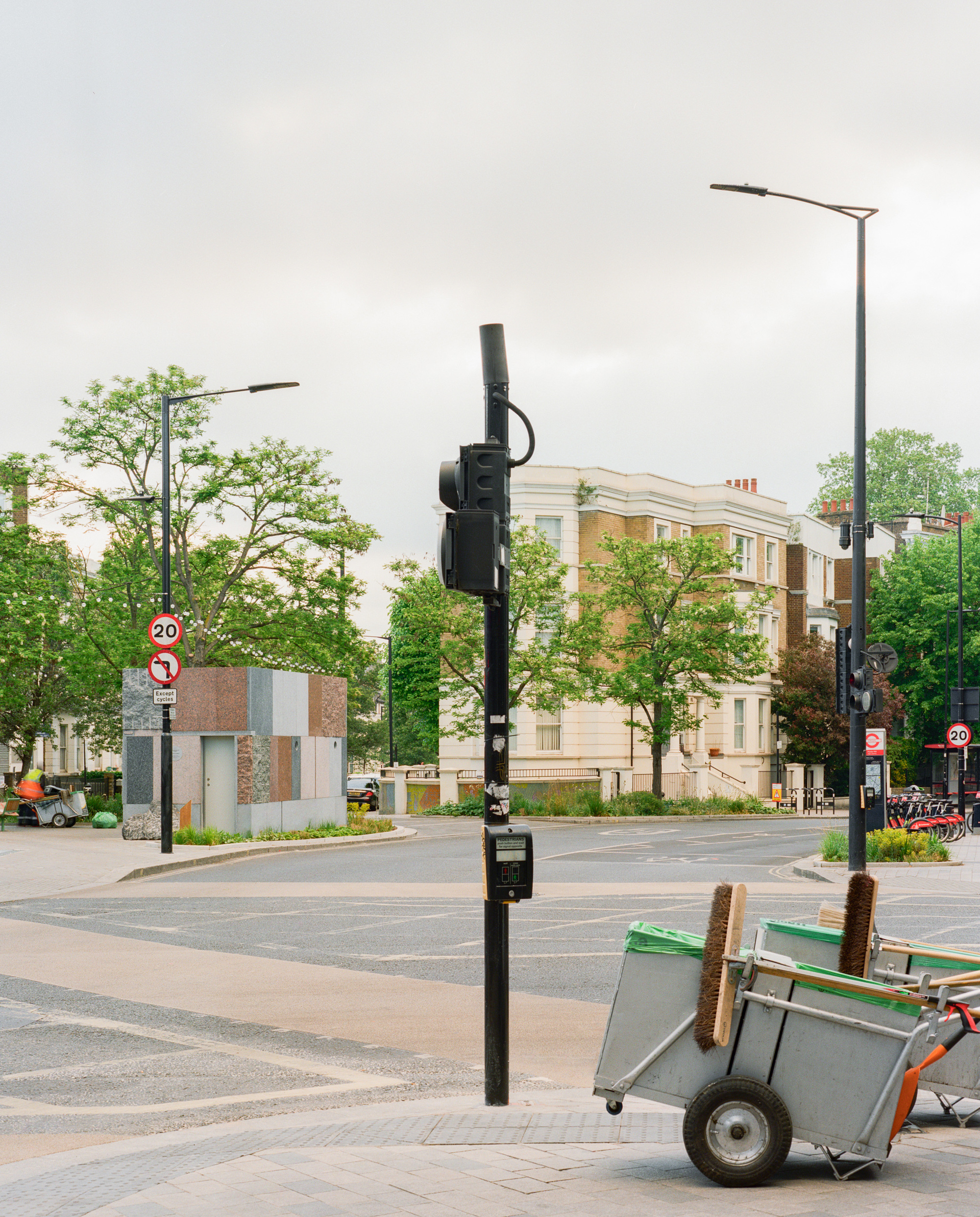 Stone-clad kiosk at a road junction beside a street cleaner's cart and traffic signal under an overcast sky