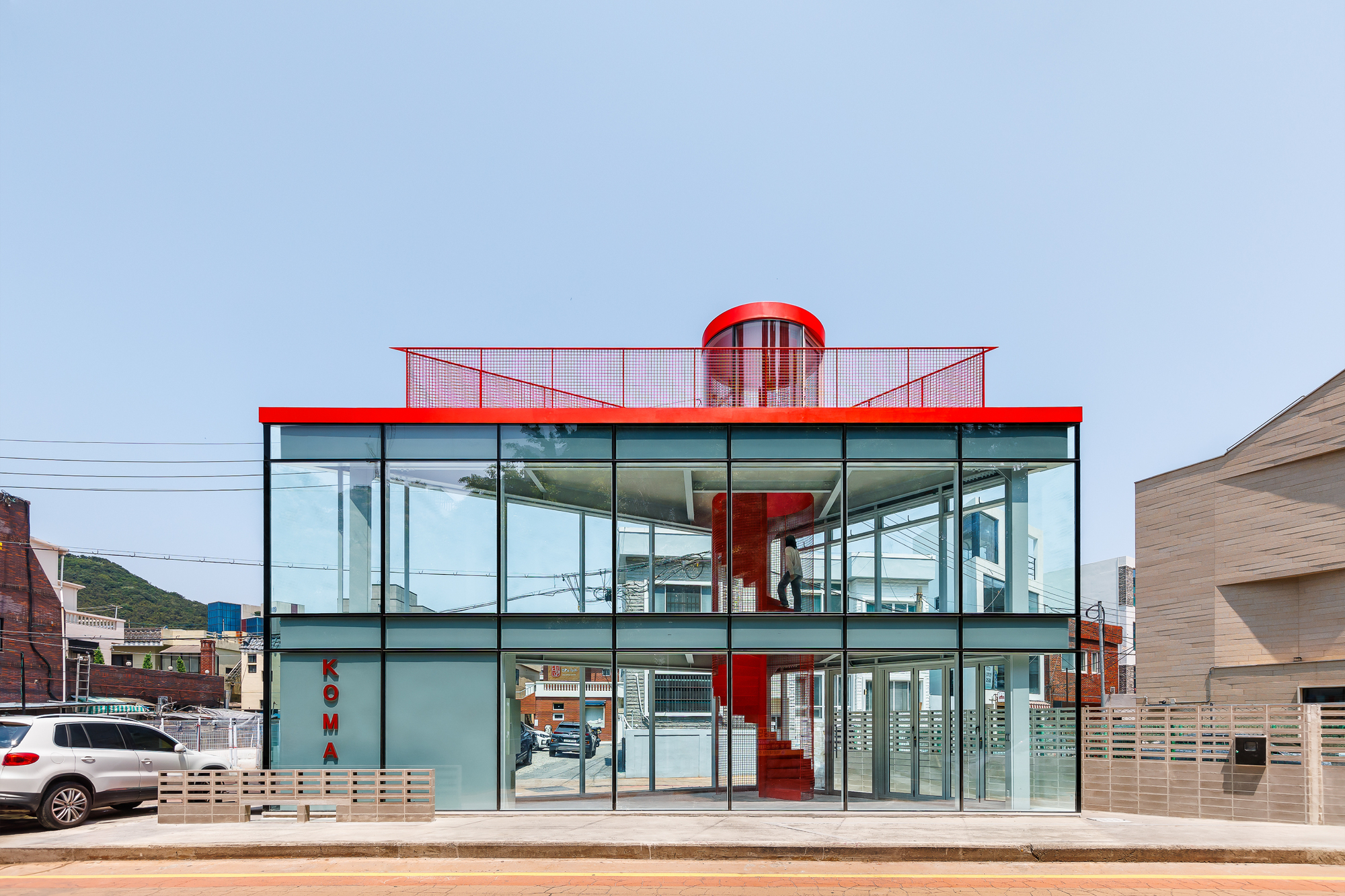 Street-level view of the glass-walled two-storey box with red roof edge, mesh parapet and cylindrical stair top under a clear sky