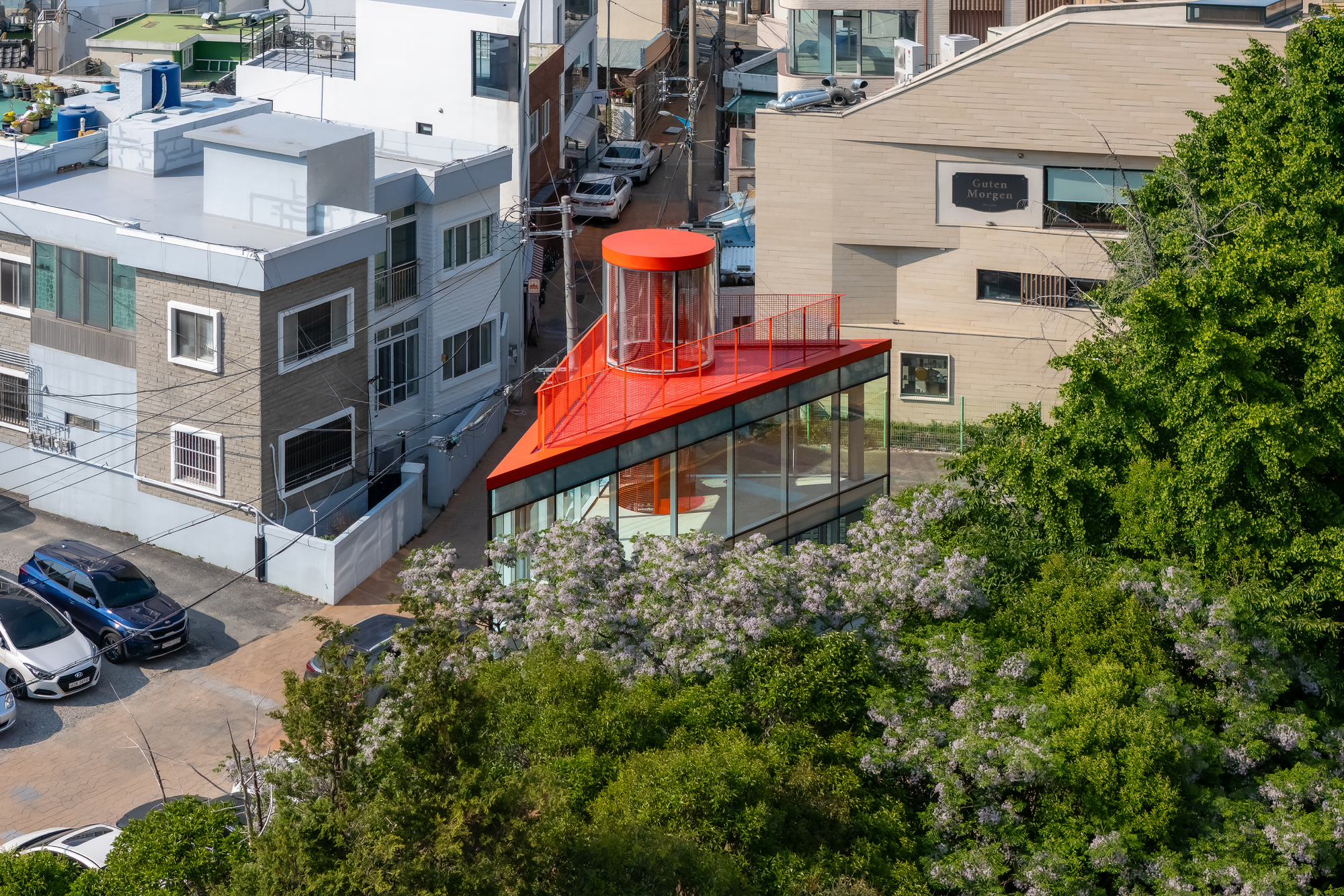 Aerial view of the red roof and glass cylinder rising above blooming lilac trees beside neighbouring low-rise houses