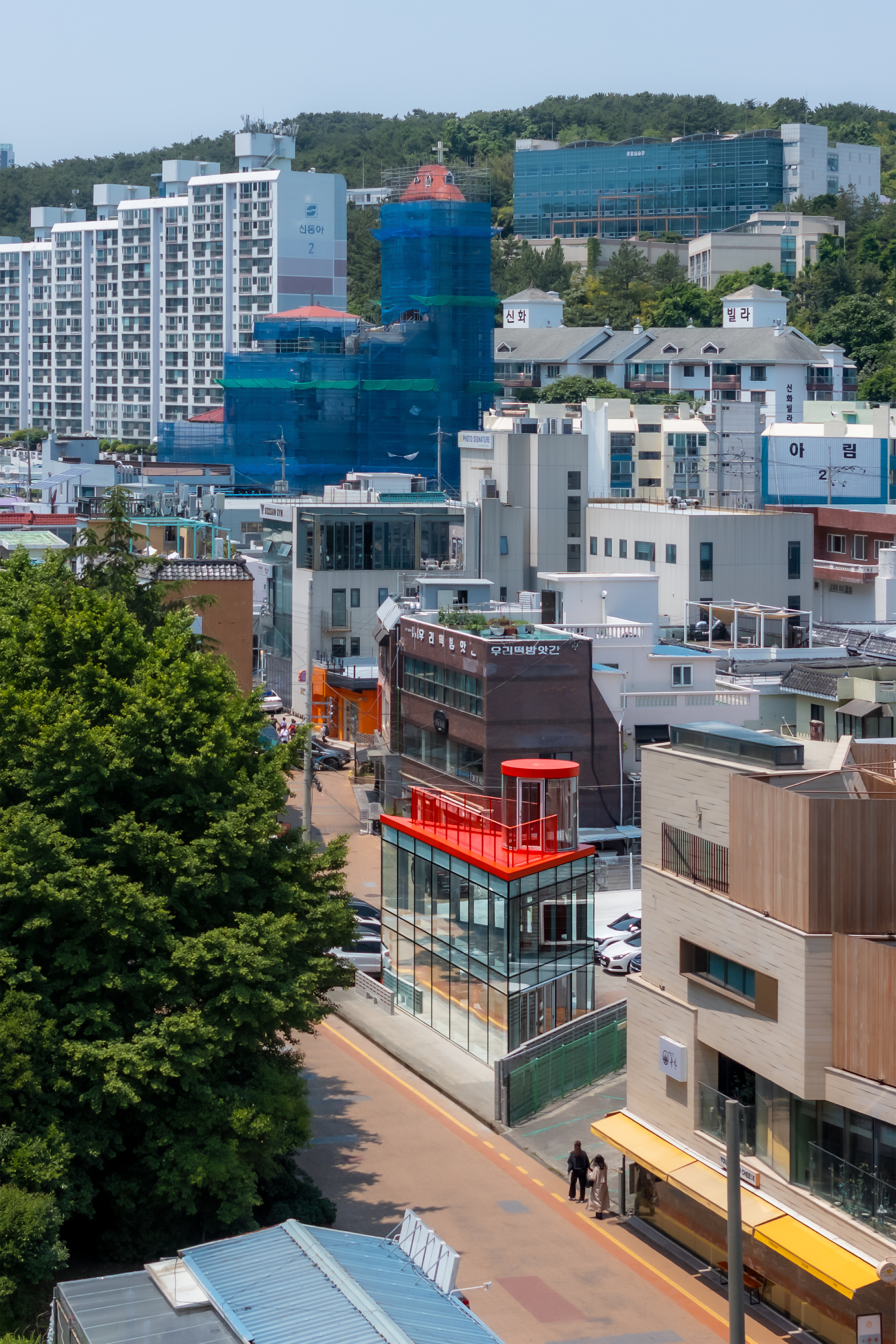 Elevated view of the red-roofed glass building on a sloping street, with pedestrians passing below and dense hillside housing behind