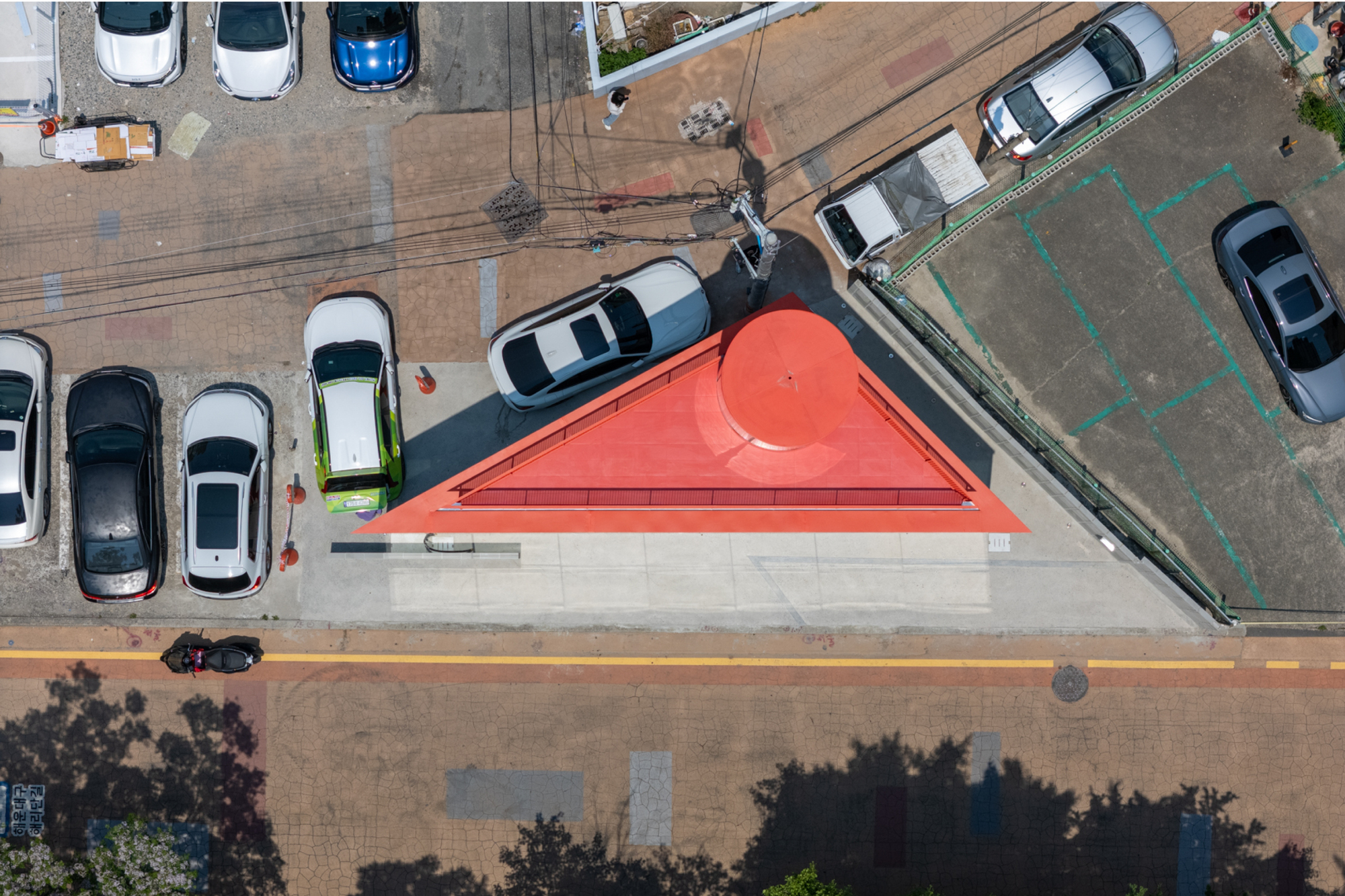 Top-down view of the triangular red roof with a round cylindrical cap, surrounded by parked cars and tree shadows