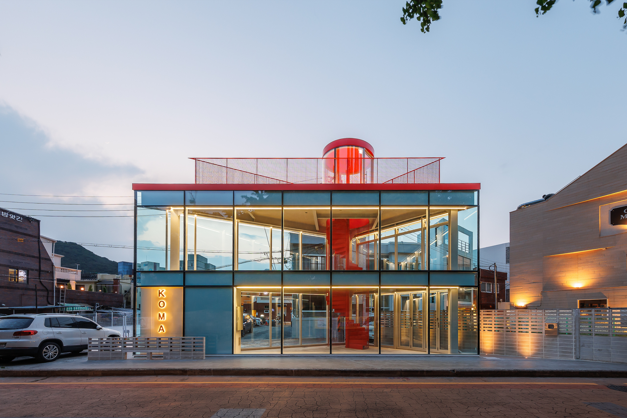 Front elevation of the two-storey glass box with red roof edge and visible red spiral stair at dusk