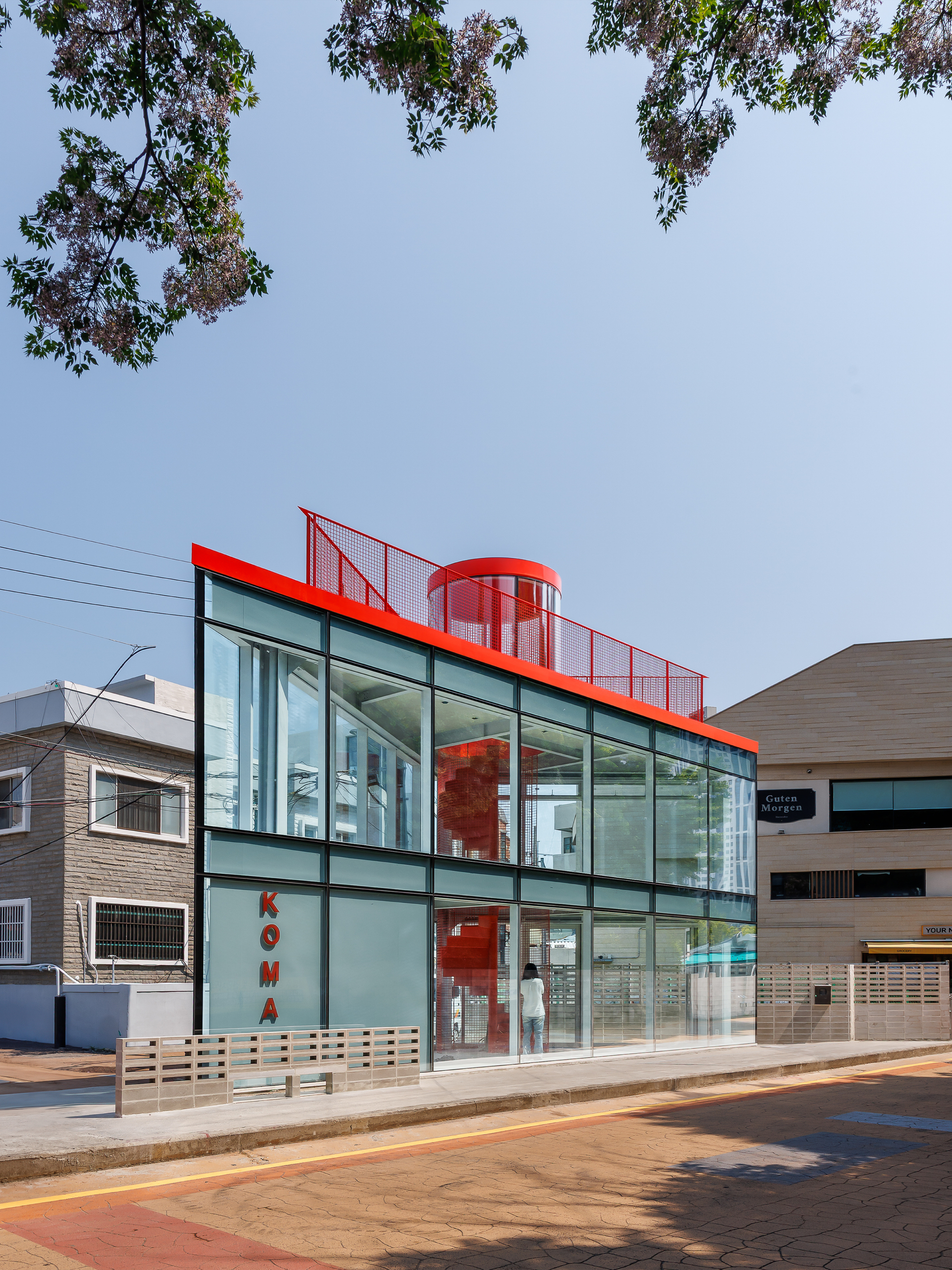 Corner view of the glazed facade with red roof fascia and mesh railing under a branch of purple blossoms