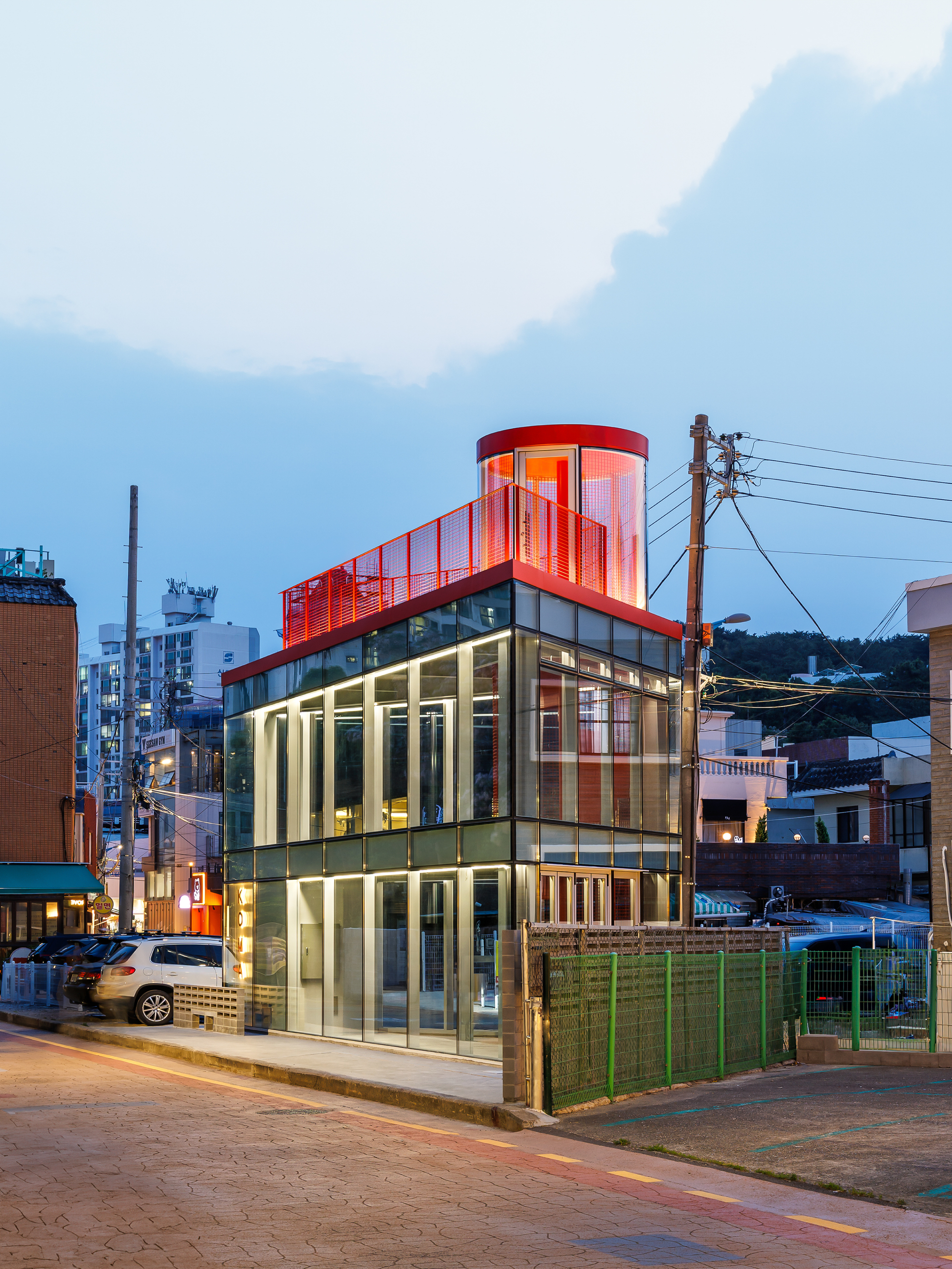Corner of the lit glass building with red cylindrical rooftop volume against a deep blue evening sky and utility poles