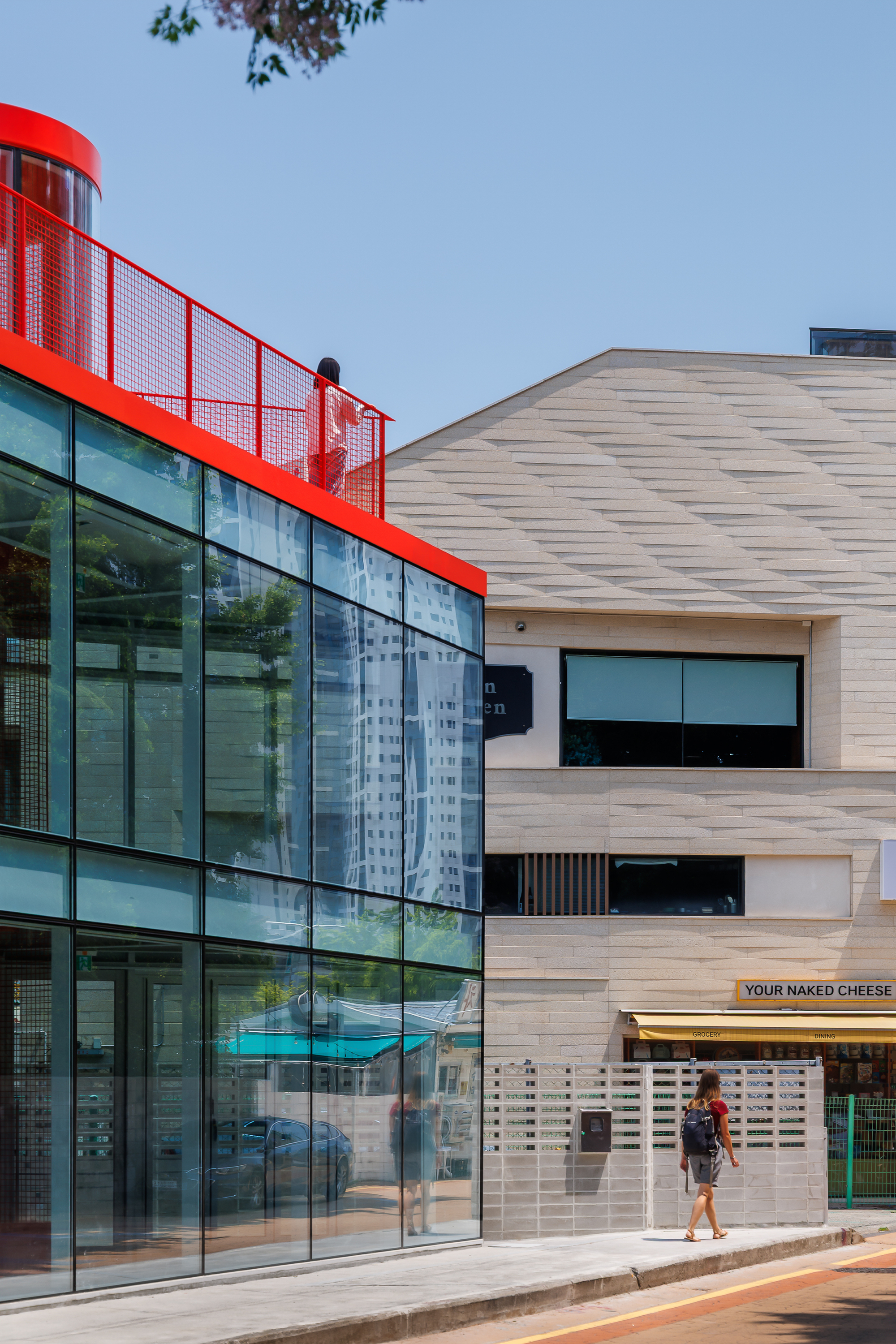 Glass facade reflecting neighboring towers beside a stone-clad building, with a pedestrian walking past and a person on the rooftop terrace