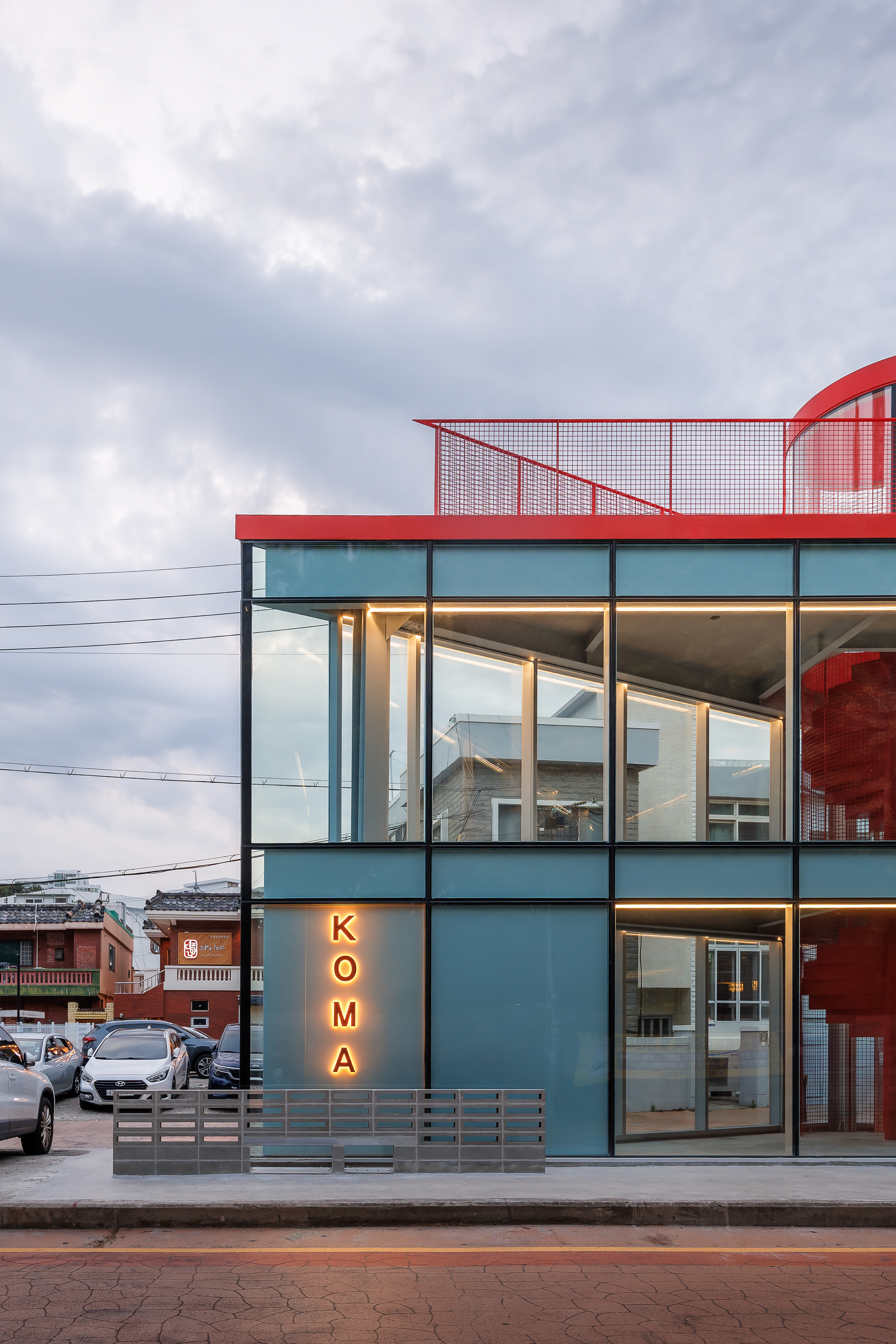 Close view of the teal panel and glass facade with glowing vertical KOMA sign under an overcast evening sky