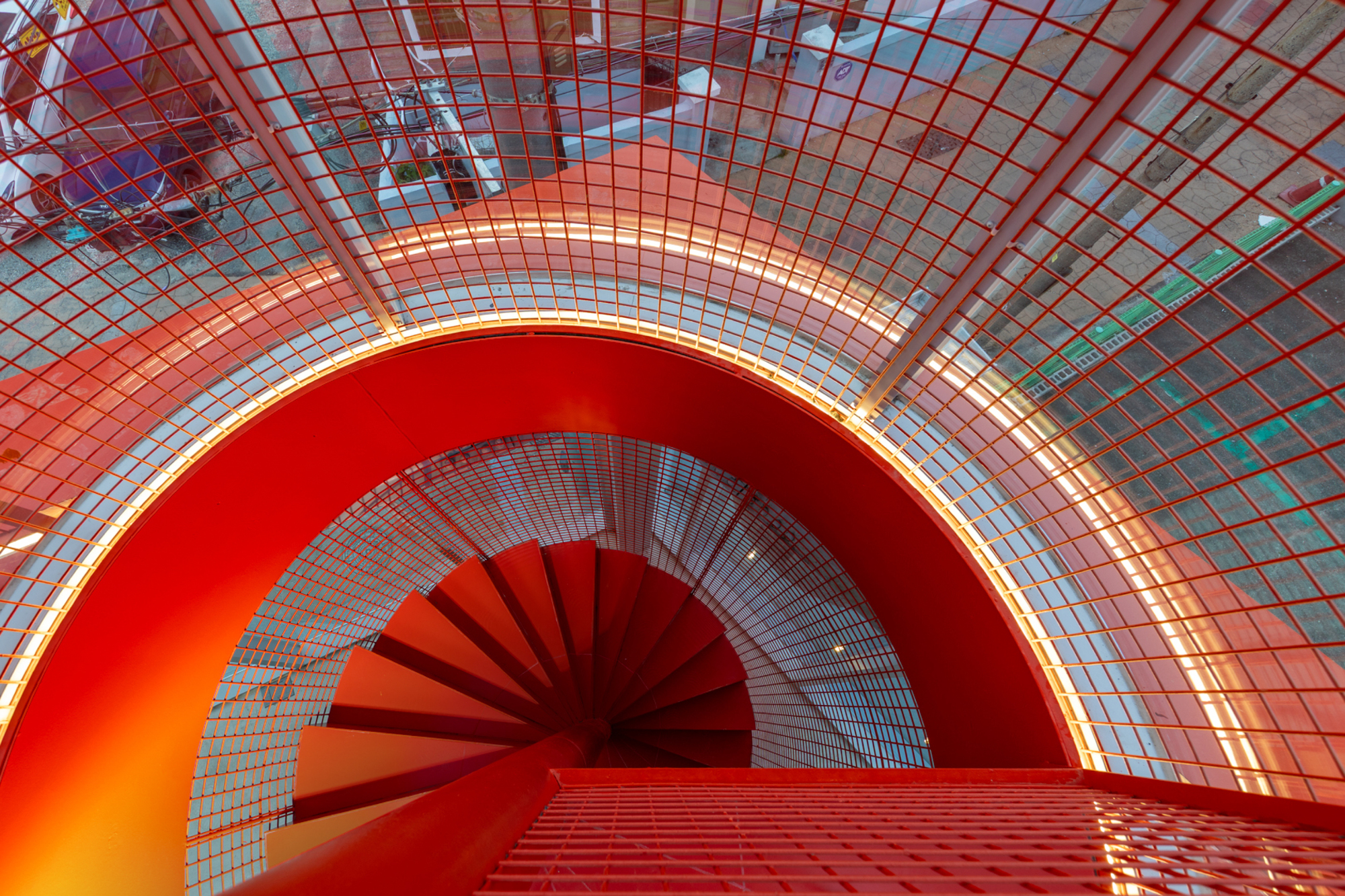 Looking down the red spiral staircase through a curved perforated metal mesh shell lit by a glowing edge strip