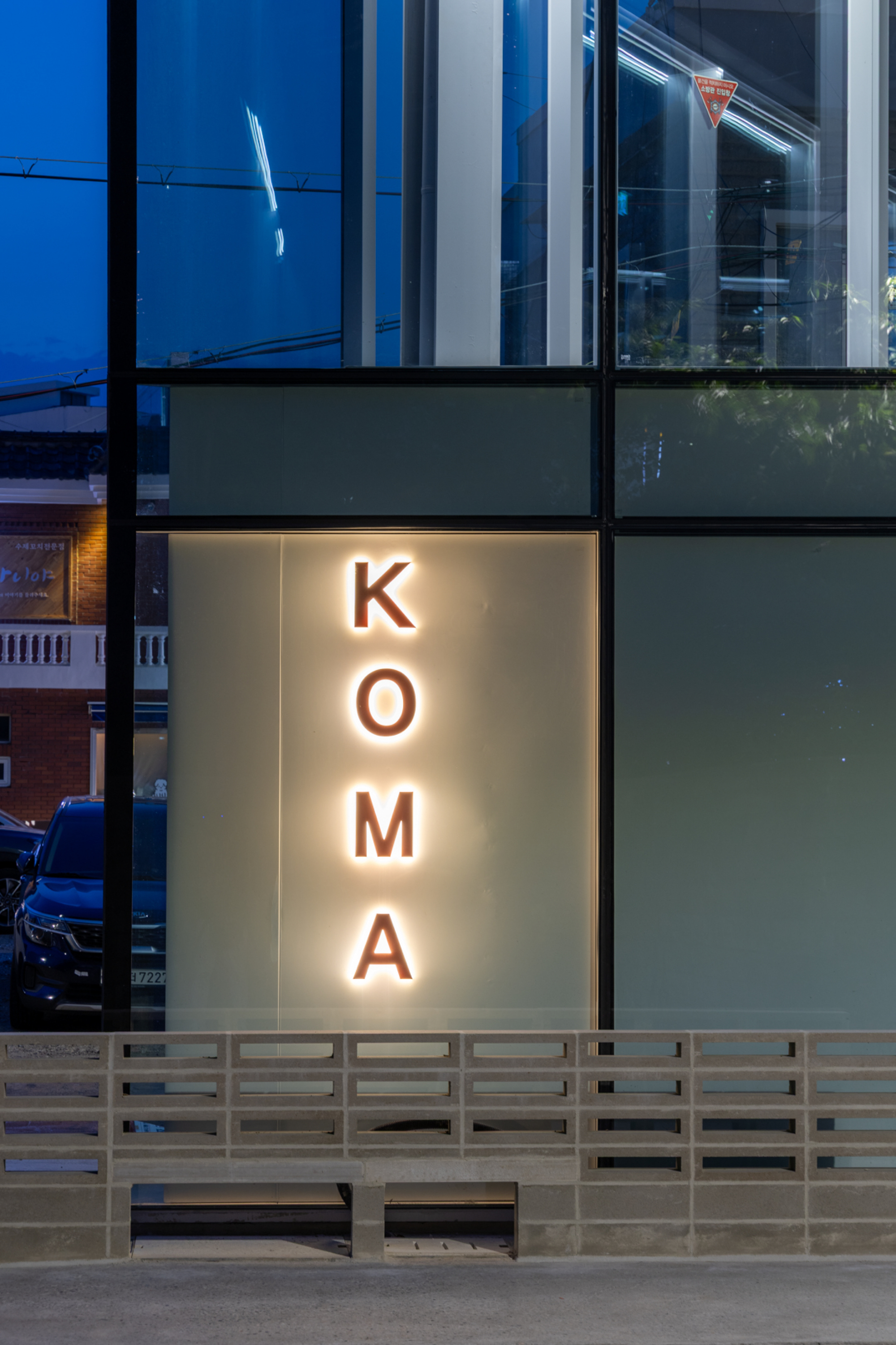 Backlit KOMA lettering on a translucent panel in a glass facade above a concrete block base at dusk