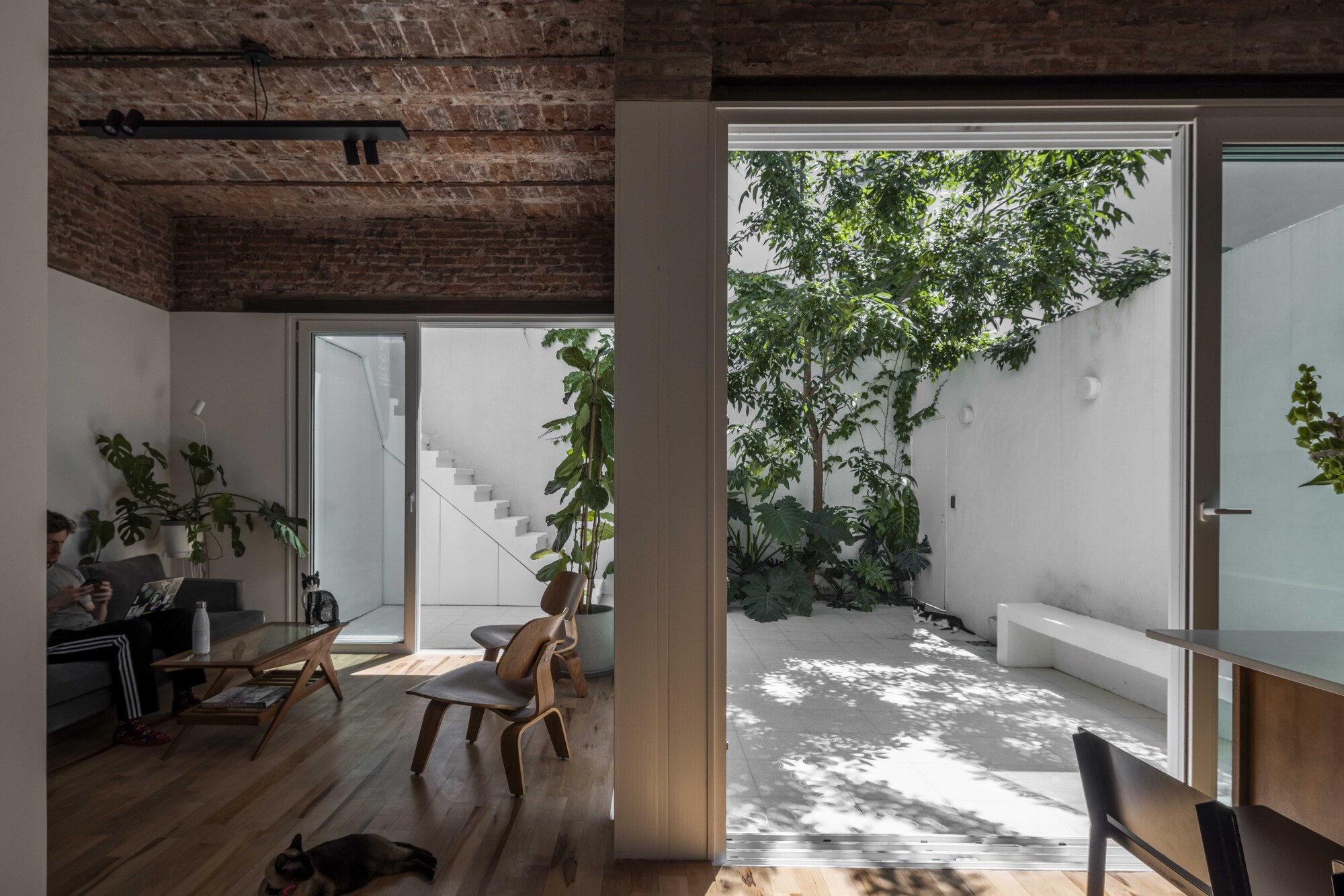 Living room with exposed brick vaulted ceiling opening through sliding glass to a planted white courtyard in afternoon sun