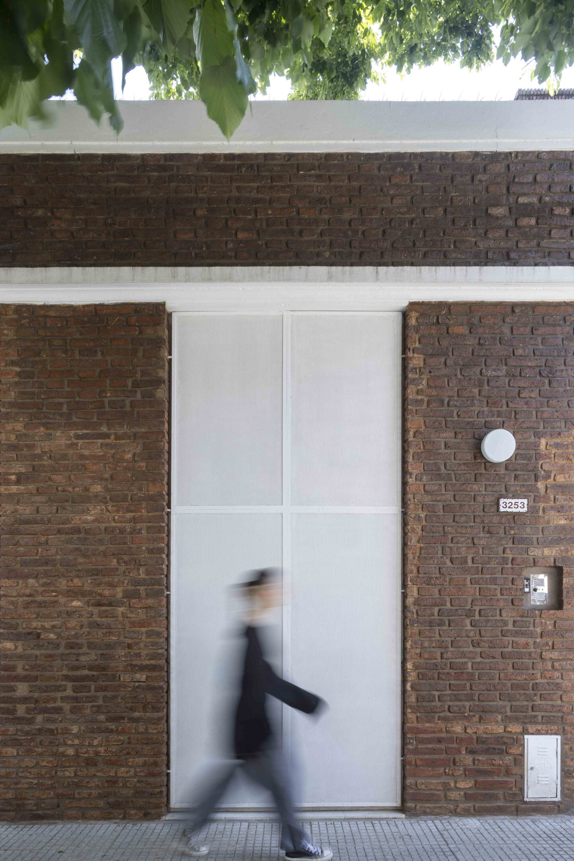 Brick street facade with a tall translucent panel door and a blurred pedestrian walking past under a tree canopy