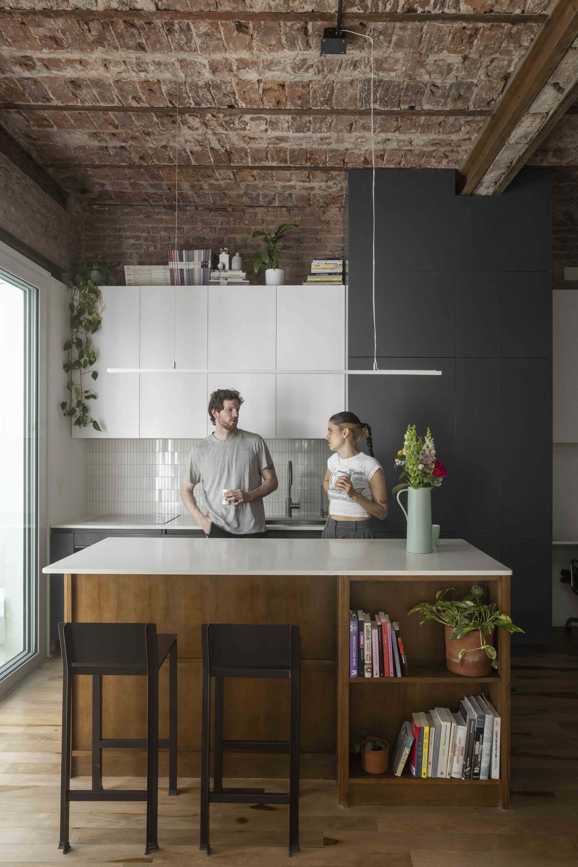 Two people stand at a timber kitchen island with open book shelves, beneath an exposed brick ceiling and black cabinetry