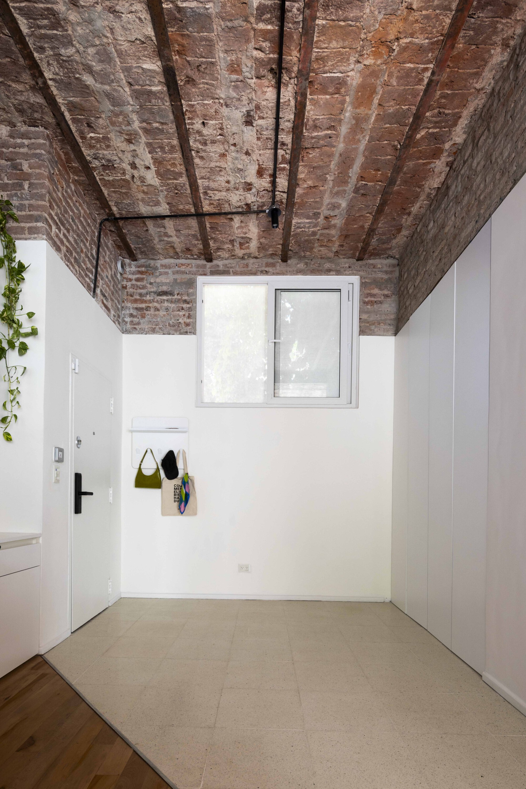 Entry hall with a vaulted exposed brick ceiling, white walls, a small window and bags hanging on a wall hook beside the door