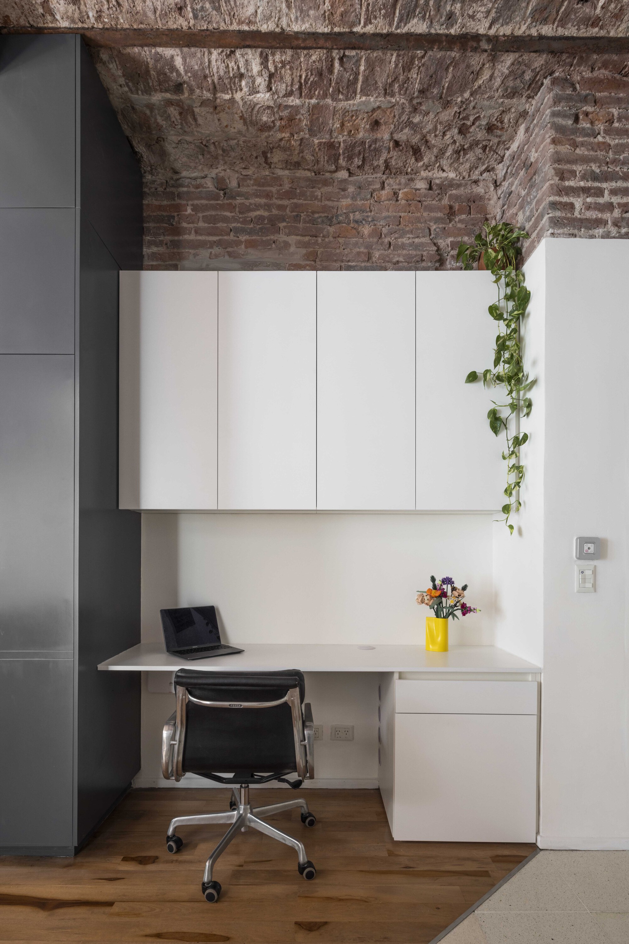 Built-in white desk with overhead cabinets and a black office chair beneath the brick vault, with a trailing plant at the side