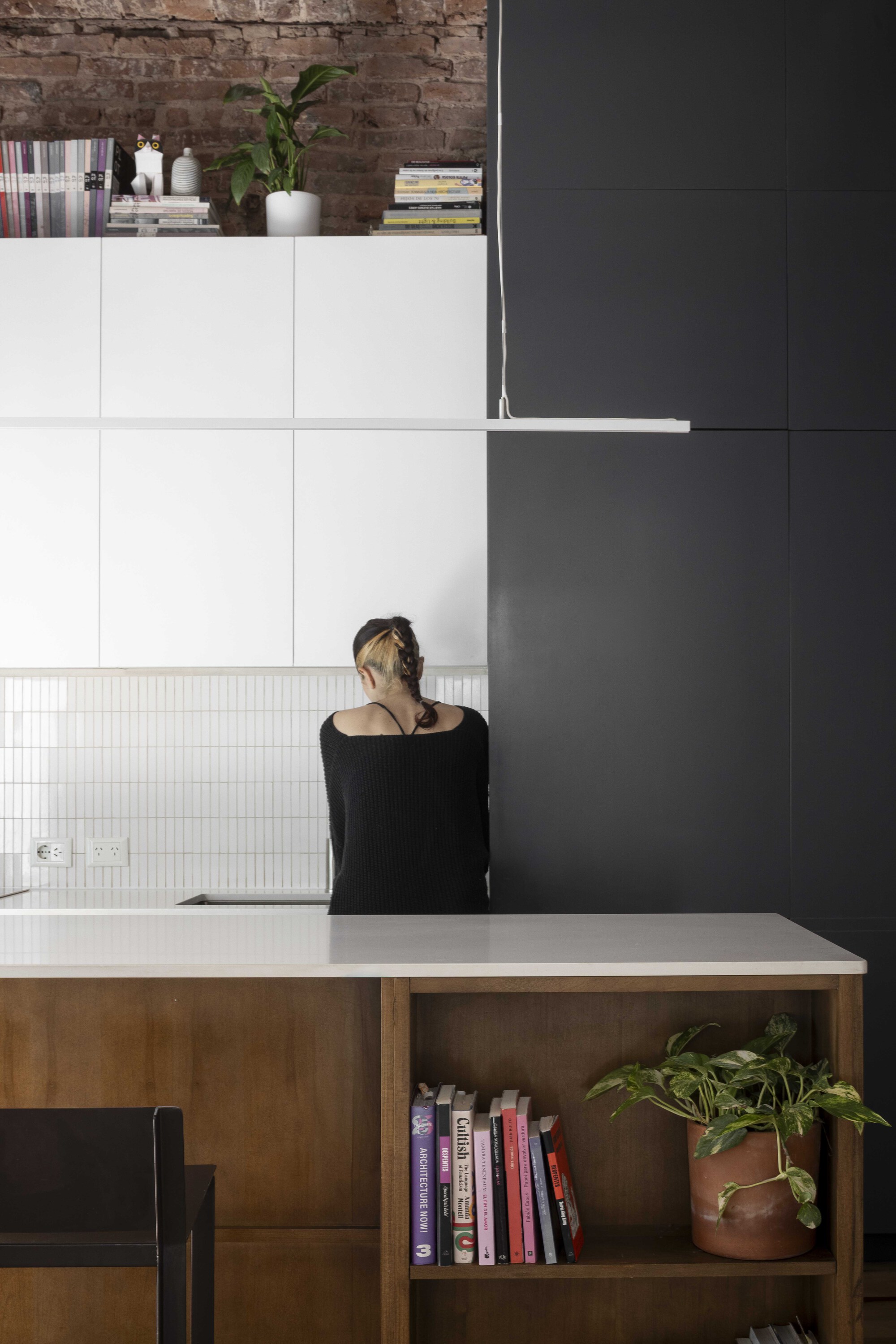 Person standing at the kitchen counter before white cabinets and a dark panel wall, with a potted plant on the bookshelf end