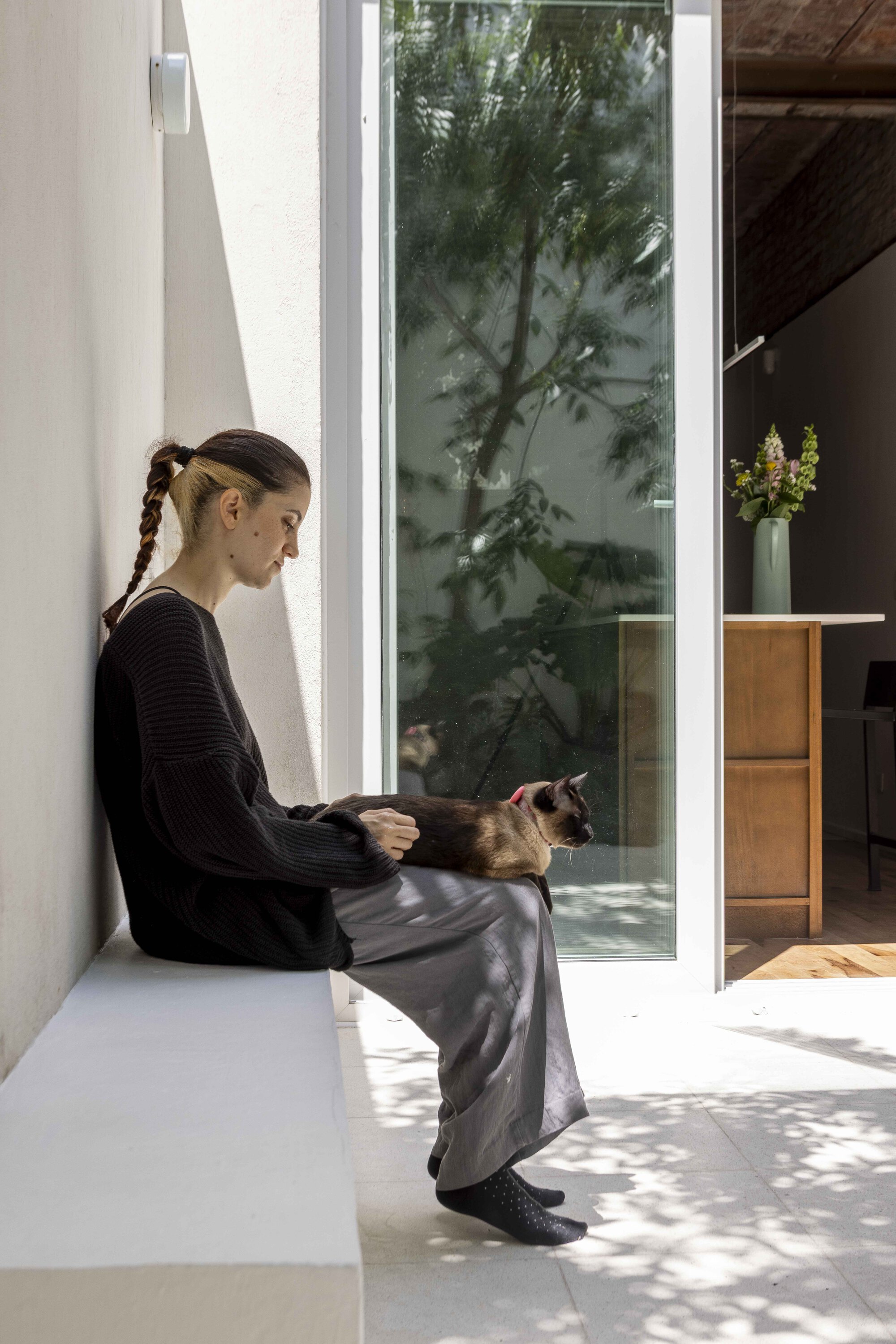 Woman with a braid sits on a white built-in bench holding a cat beside a glass door in dappled sun