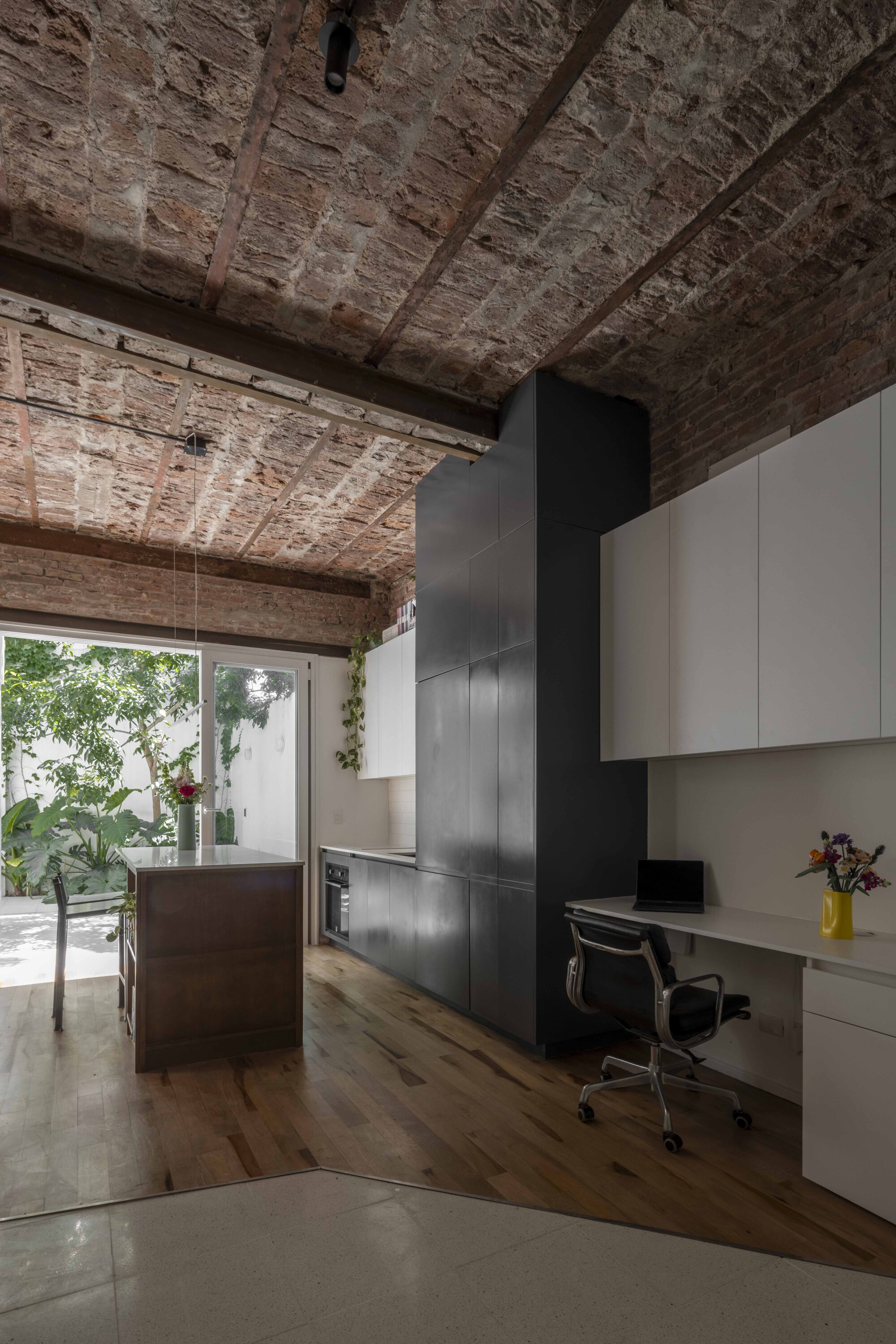 Interior with exposed brick vaulted ceiling, dark cabinet tower, built-in desk and kitchen island opening to a sunlit courtyard